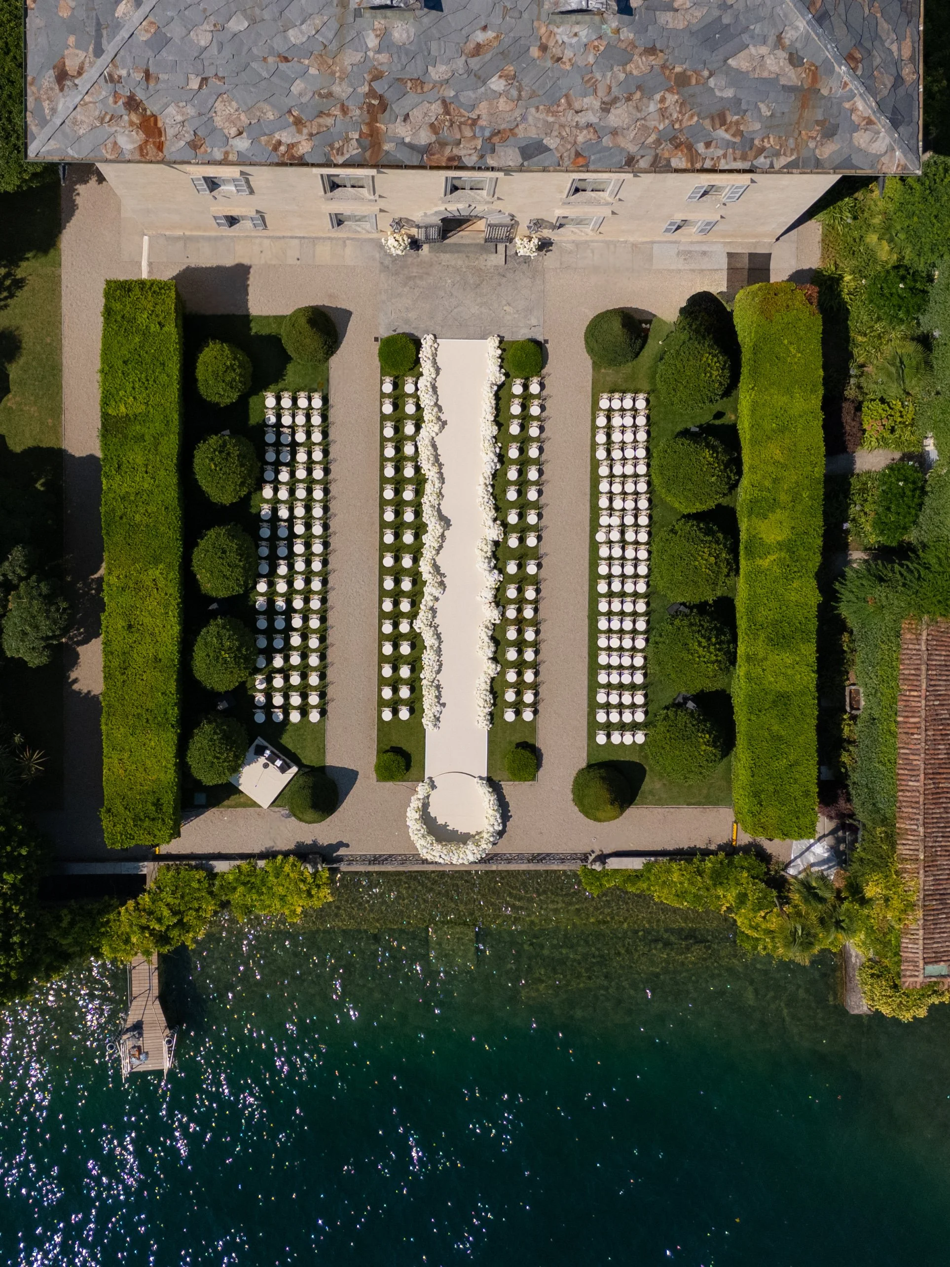 Aerial view of an outdoor wedding setup with rows of chairs and a white aisle leading to a circular floral arch, adjacent to a body of water surrounded by greenery and a stone building at the top.