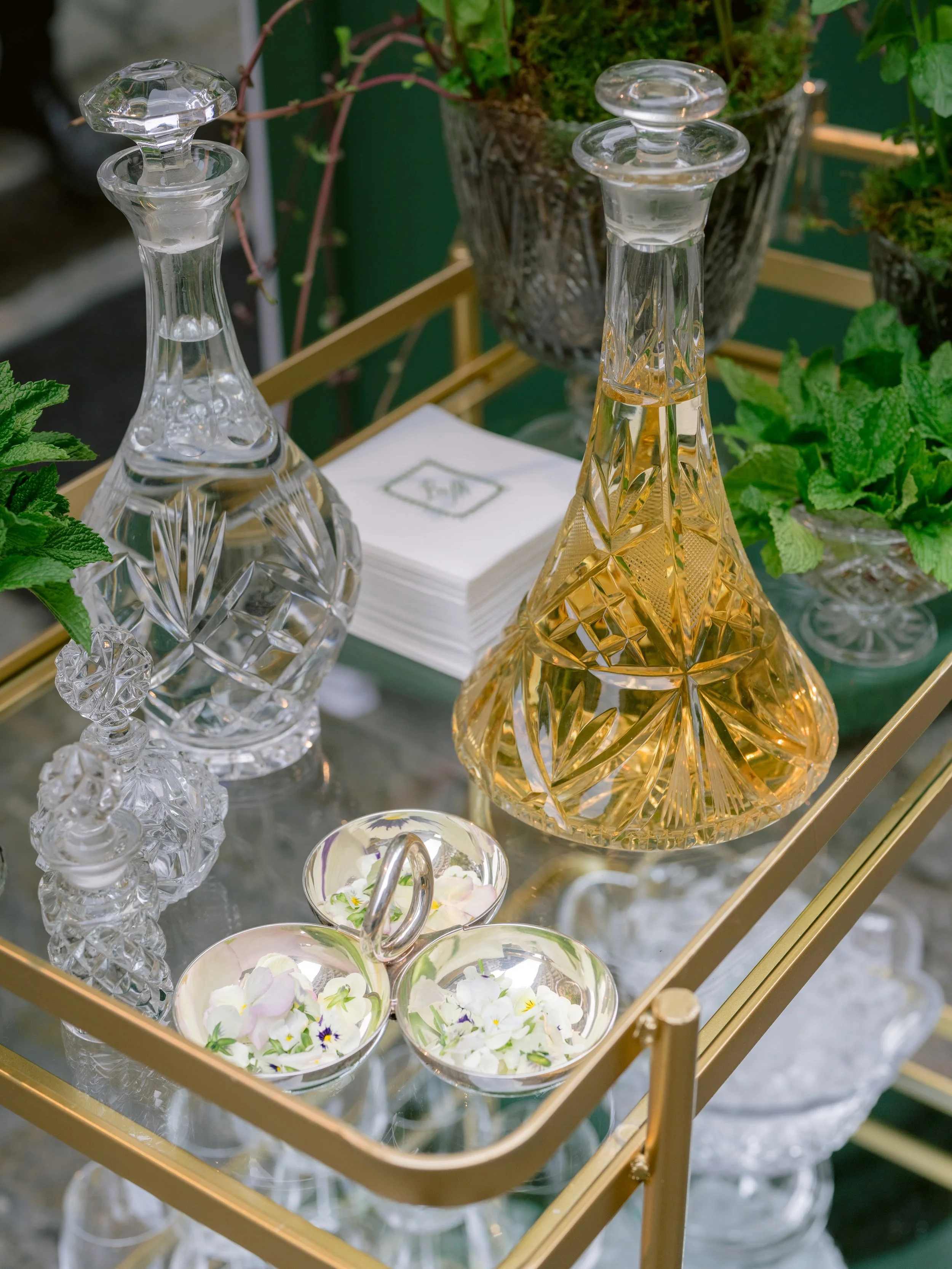 Decorative glass decanters and bowls with flowers on a glass-top cart, with potted plants in the background.