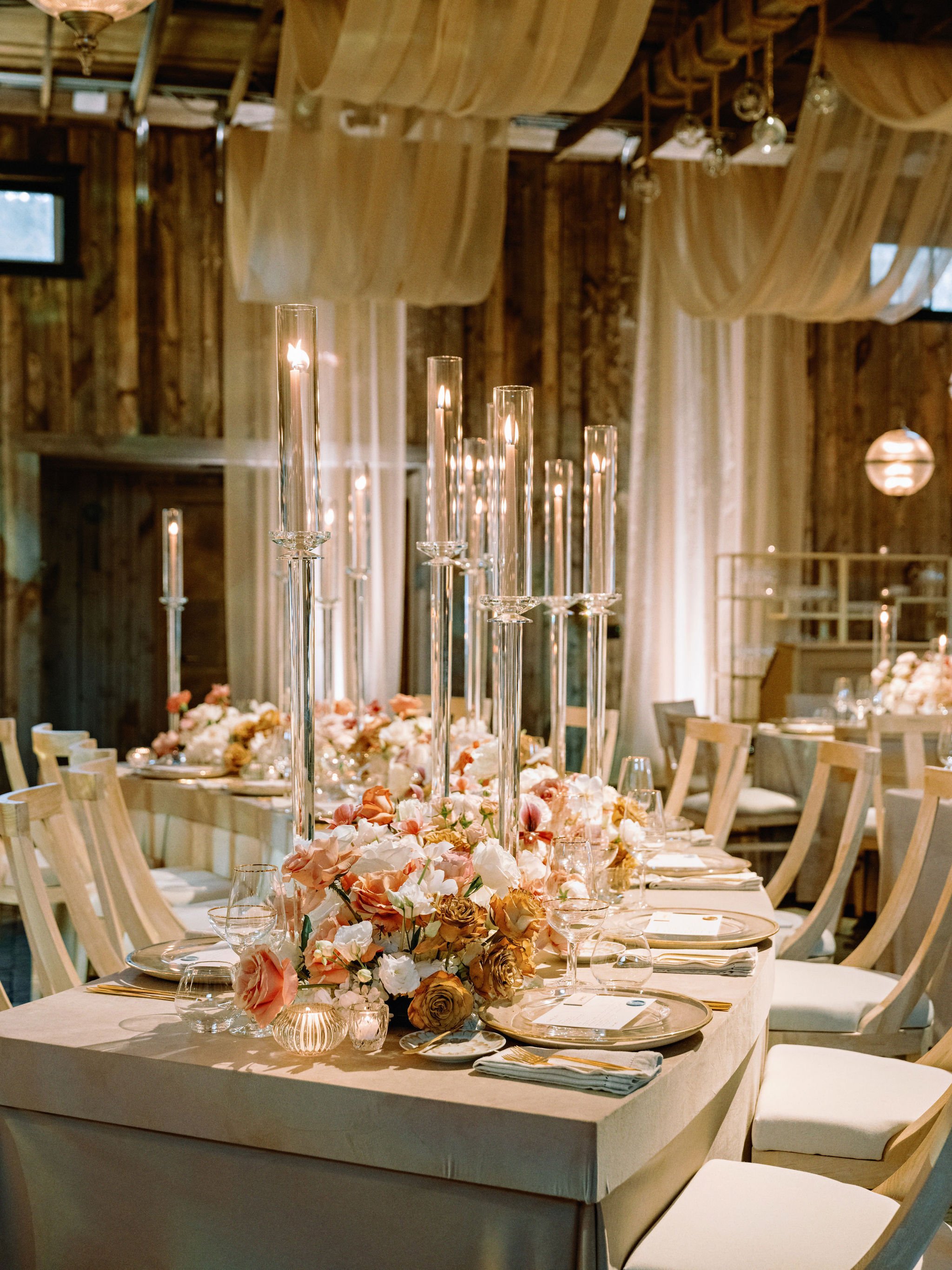 Elegant wedding reception table with floral centerpieces and tall candleholders in a rustic wooden venue with draped fabric ceilings.