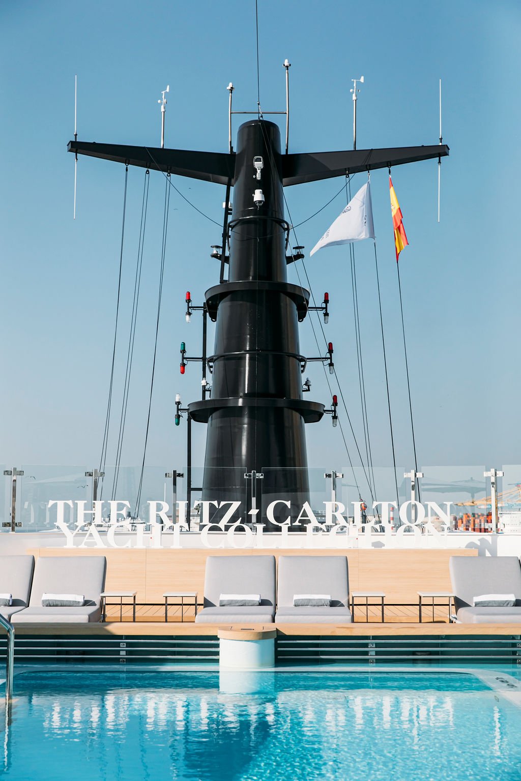 Deck with pool and lounge chairs in front of a boat with antennas, flags, and a large black mast at The Ritz-Carlton Yacht Collection.