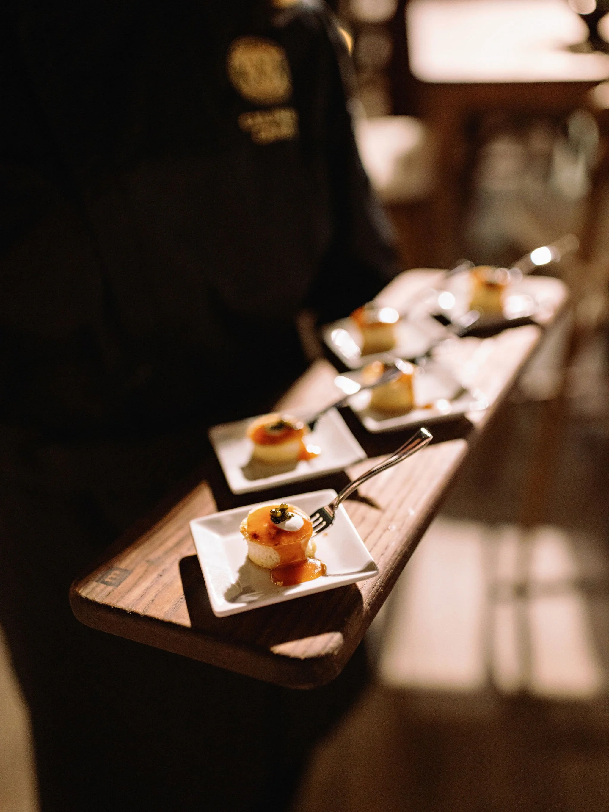 A person holding a tray of small plated appetizers on a wooden board with dim lighting.