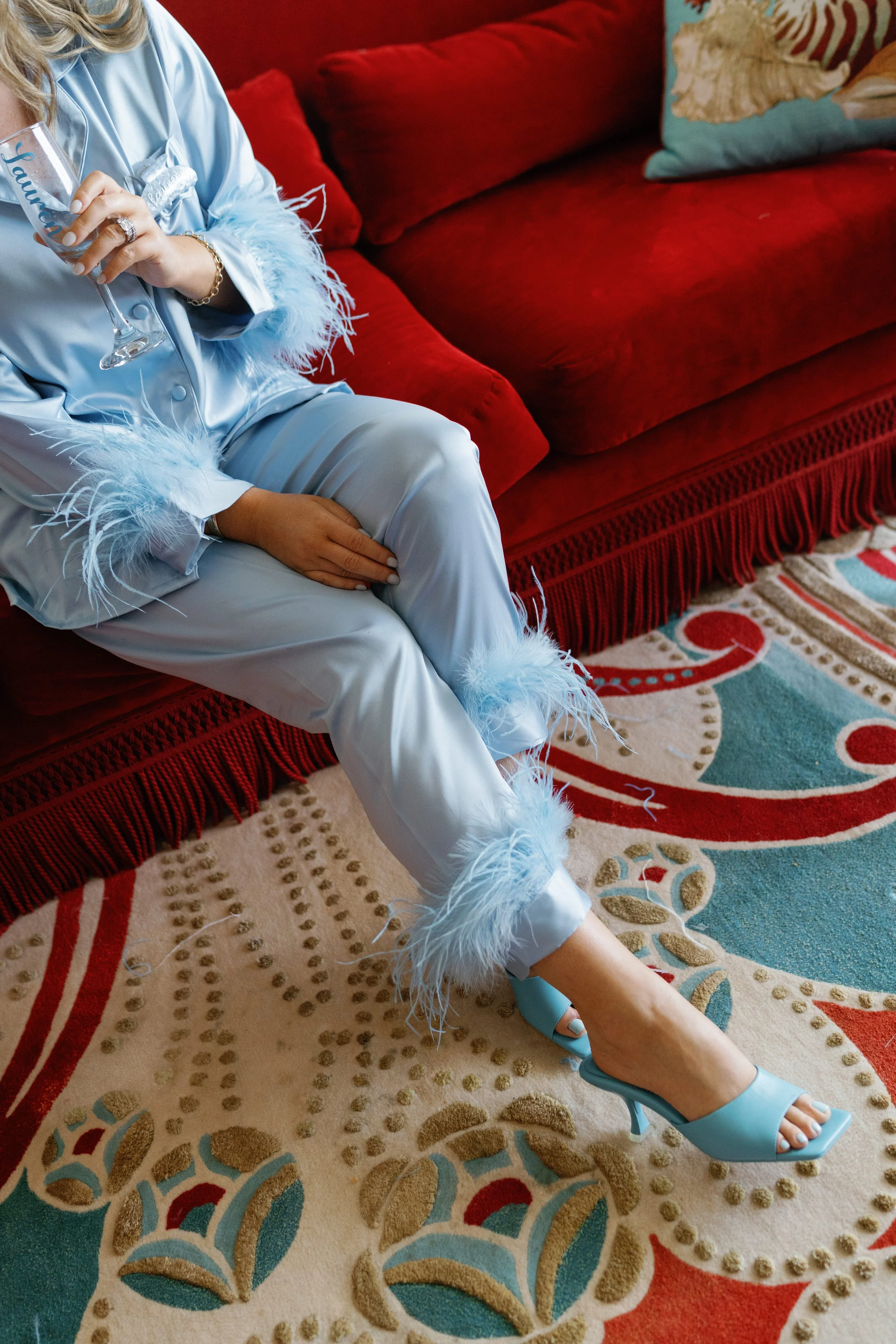 A person dressed in light blue satin pajamas with feather trim, sitting on a red velvet couch, holding a wine glass and wearing blue high-heeled shoes. An ornate patterned rug is visible on the floor.