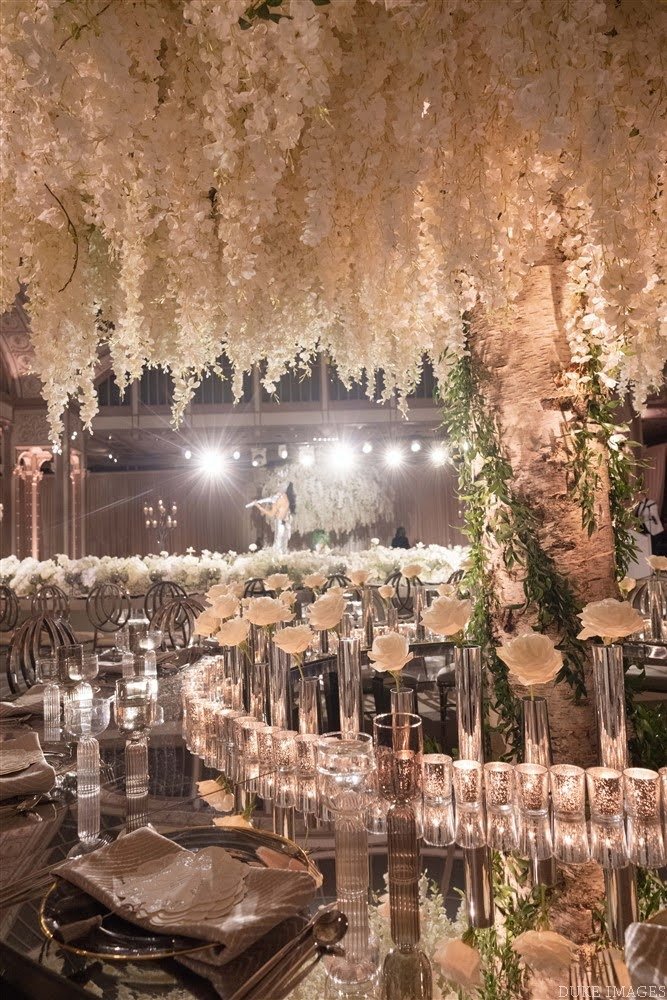 Elegant banquet hall decorated with cascading white flowers, candles, and greenery, with a stage in the background illuminated by bright lights.