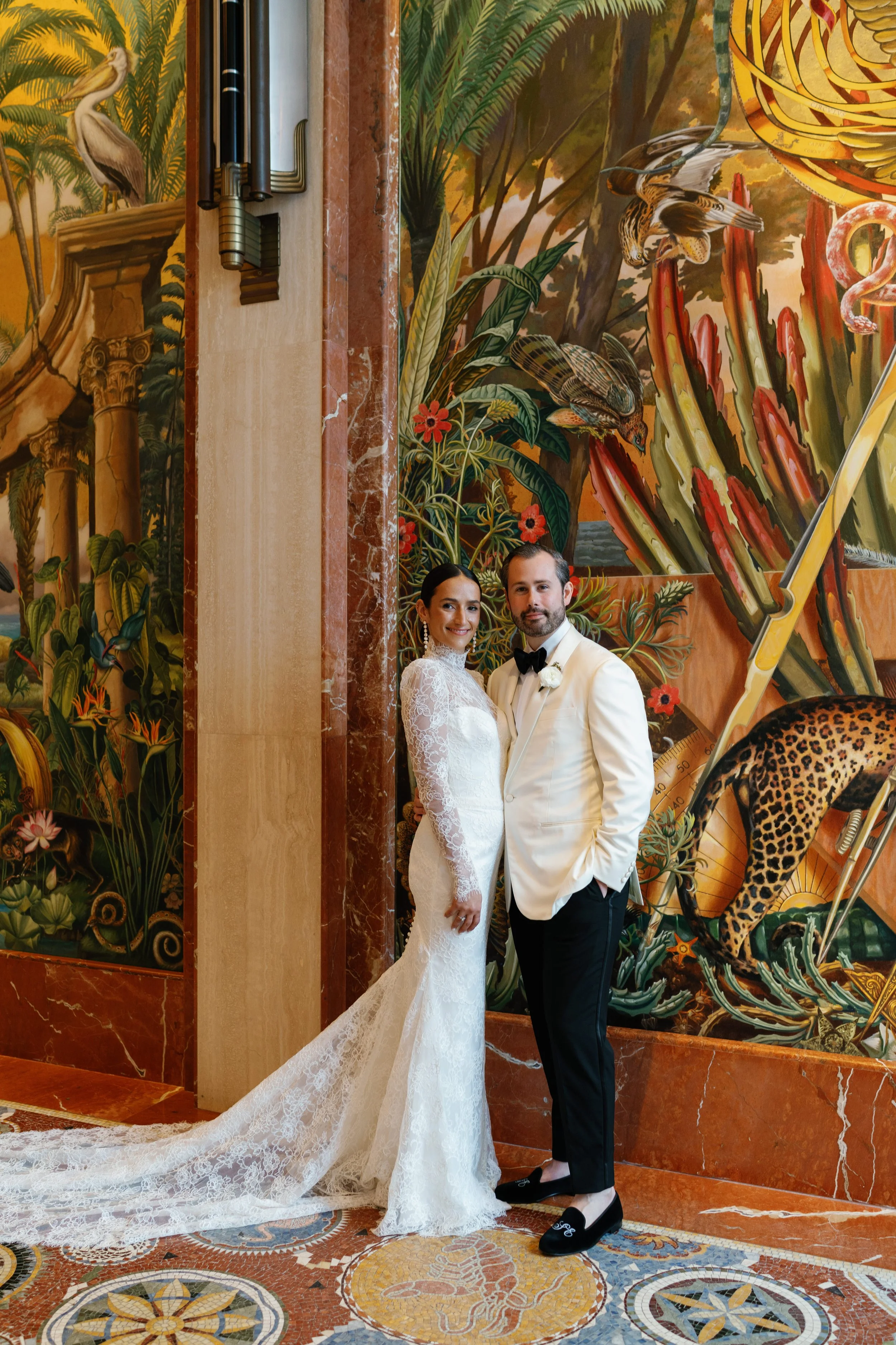 A bride and groom pose in front of a colorful jungle-themed mural at their wedding. The bride wears a white lace wedding gown, and the groom wears a white tuxedo jacket with black pants and shoes.
