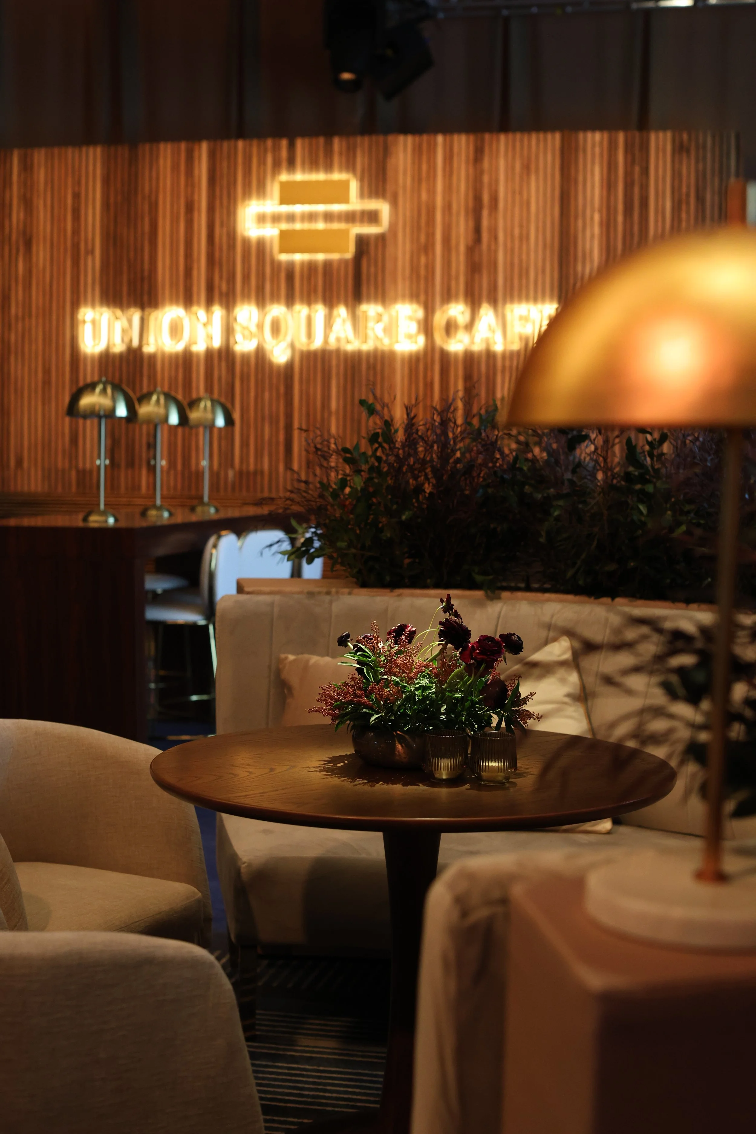 Interior of Union Square Café with a round wooden table decorated with flowers, beige chairs, and warm lighting, including a glowing neon sign with a logo and the restaurant name in the background.