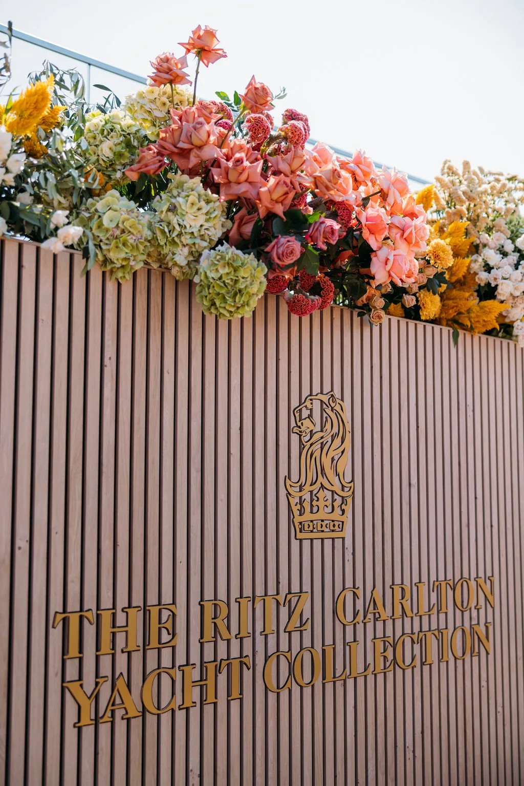 Wooden exterior wall with logo and text reading "The Ritz Carlton Yacht Collection," topped with an arrangement of pink, peach, yellow, and white flowers.