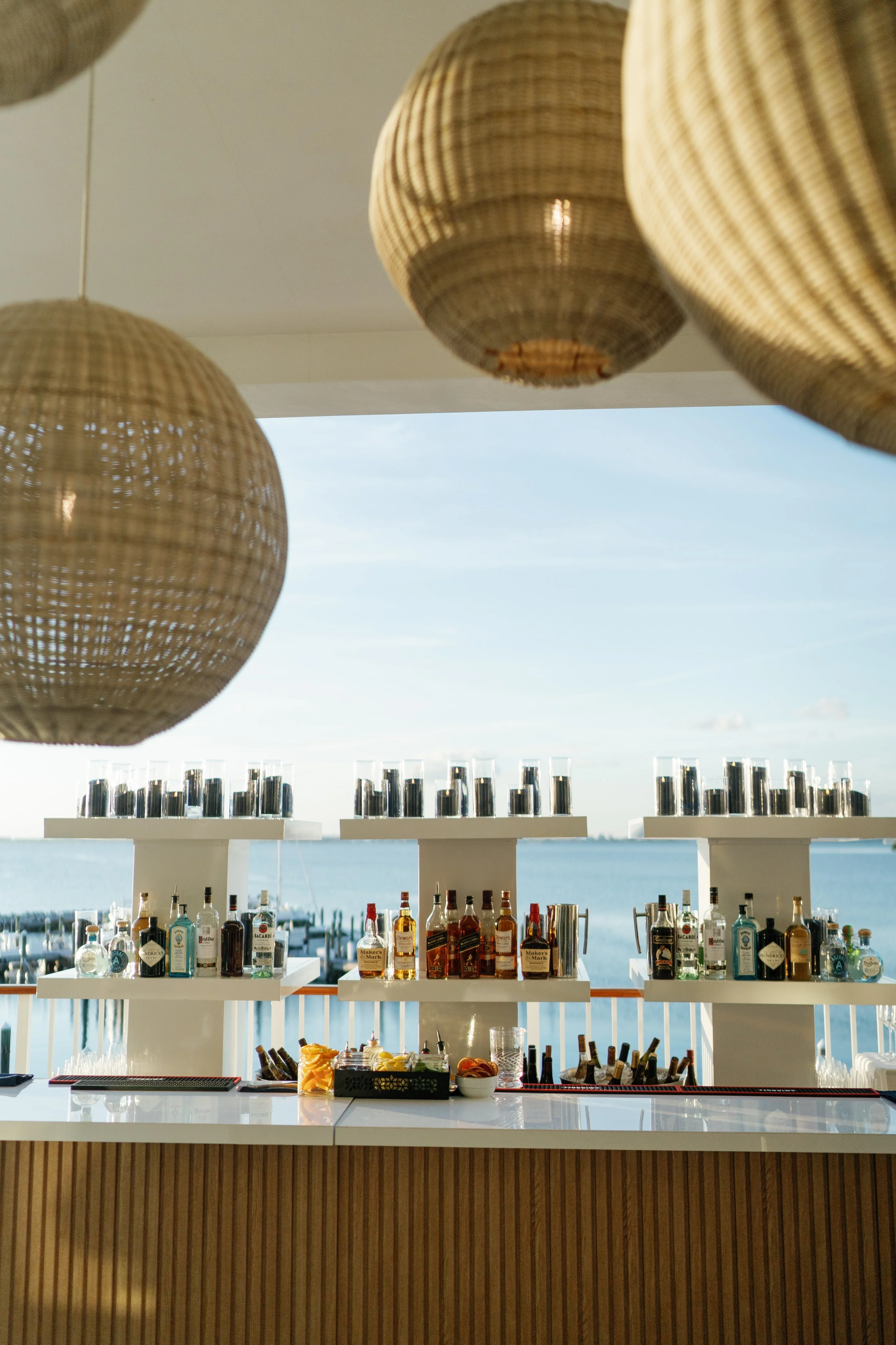 Outdoor bar setup with liquor bottles and glasses, overlooking a body of water, with round woven lanterns hanging above.