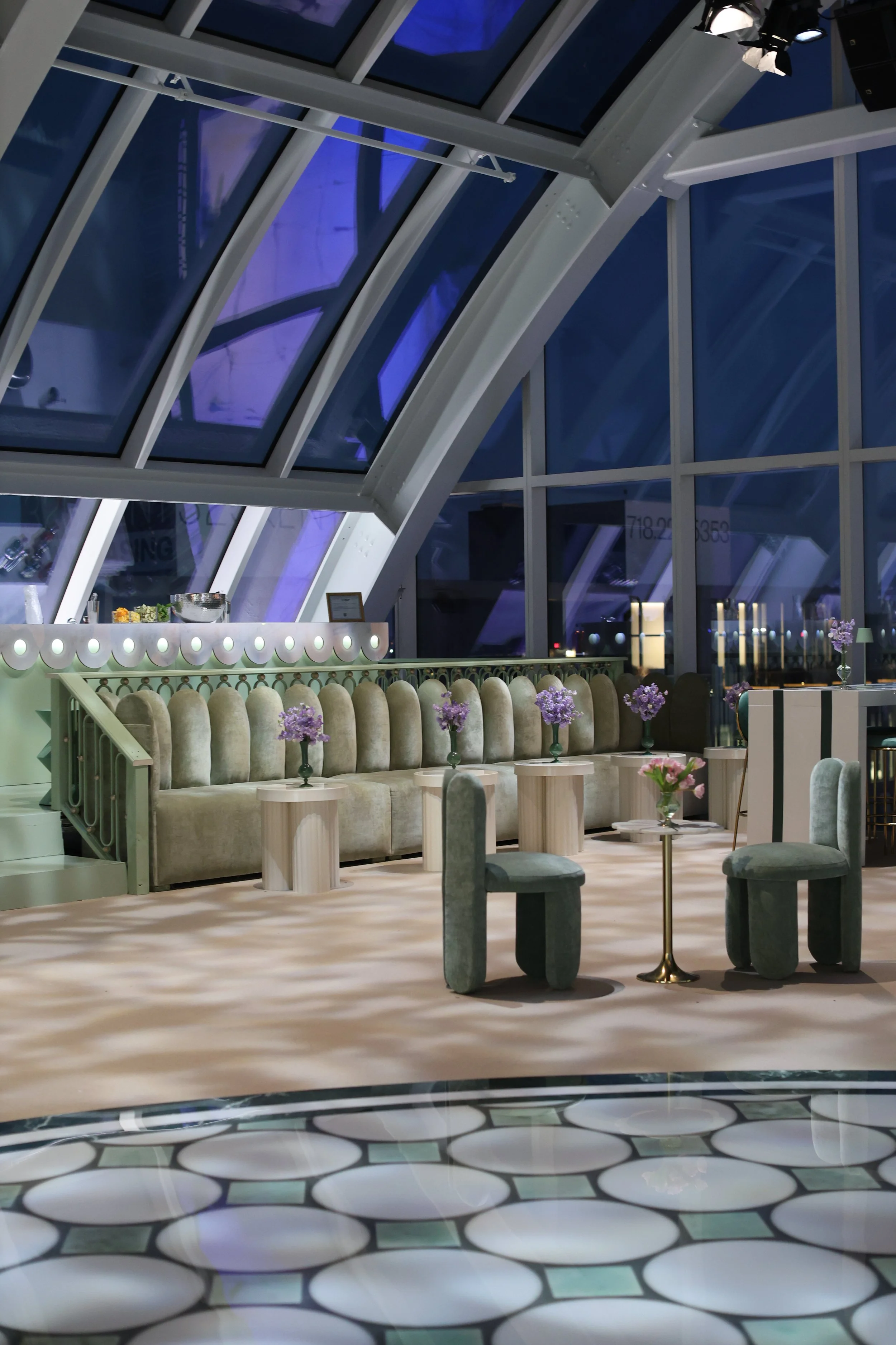 Stylish lounge area with upholstered seating, small flower vases, tables, and a glass-encased arched ceiling in a modern interior design setting.