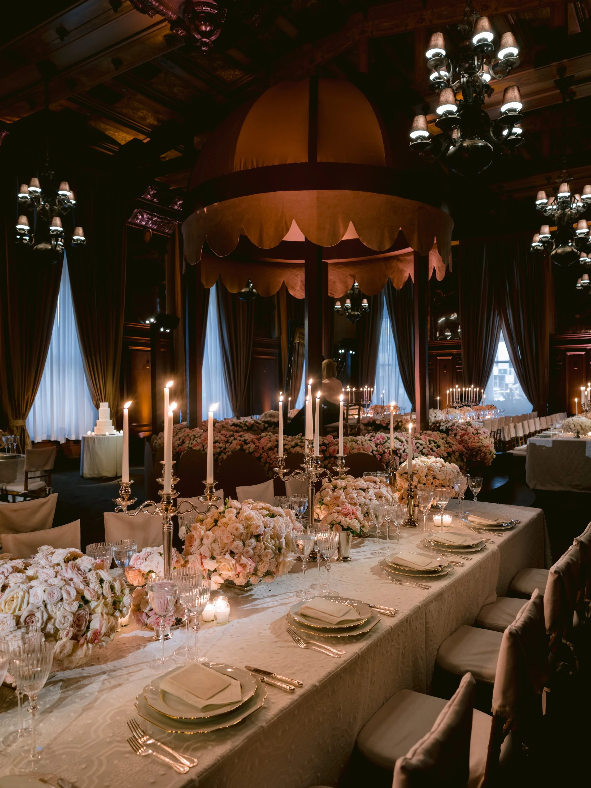 Elegant banquet hall decorated with pink and white floral arrangements, tall candles, and chandeliers, with a large canopy over the main table.