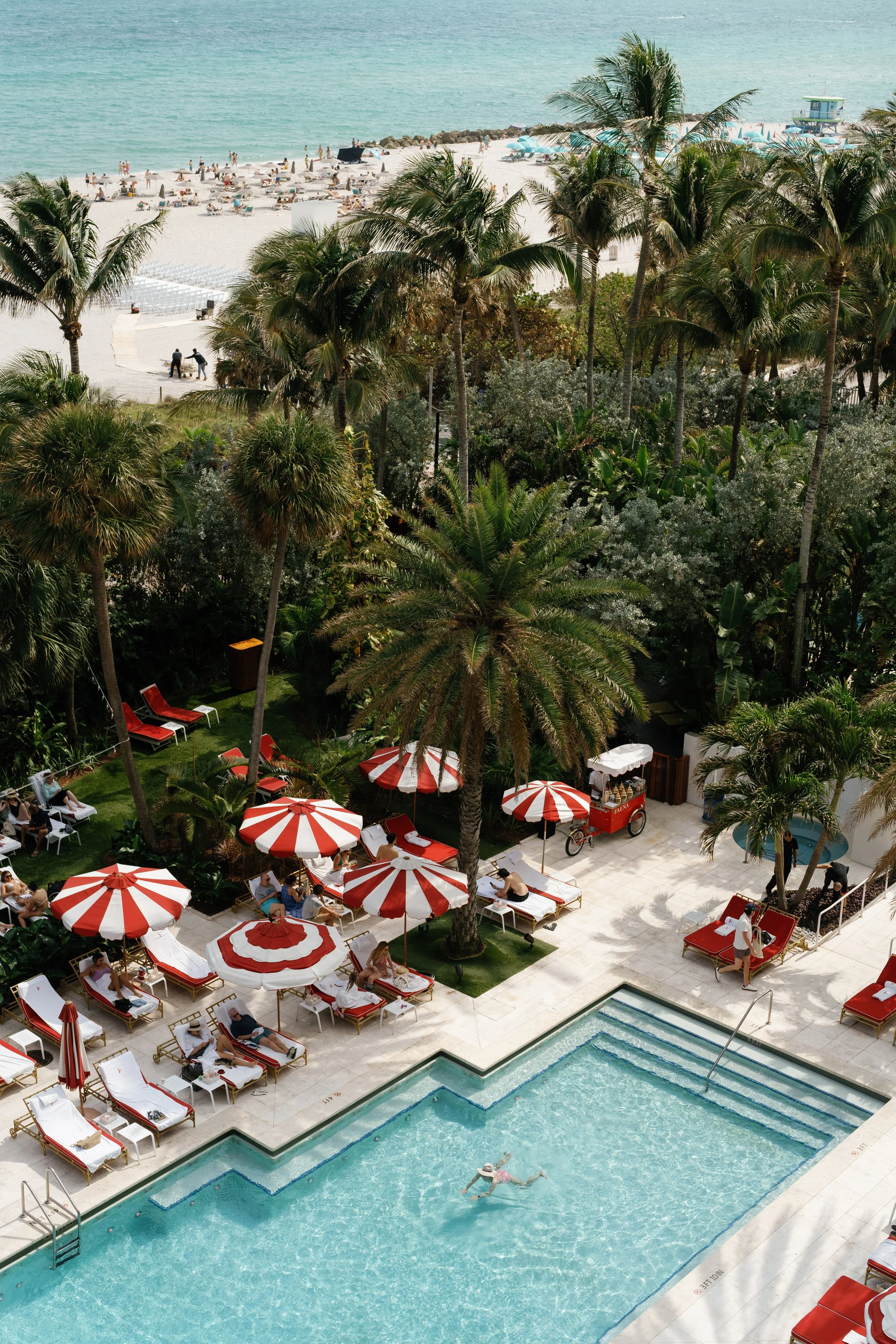 Swimming pool area with red and white striped umbrellas, lounge chairs, and a person swimming, surrounded by palm trees.