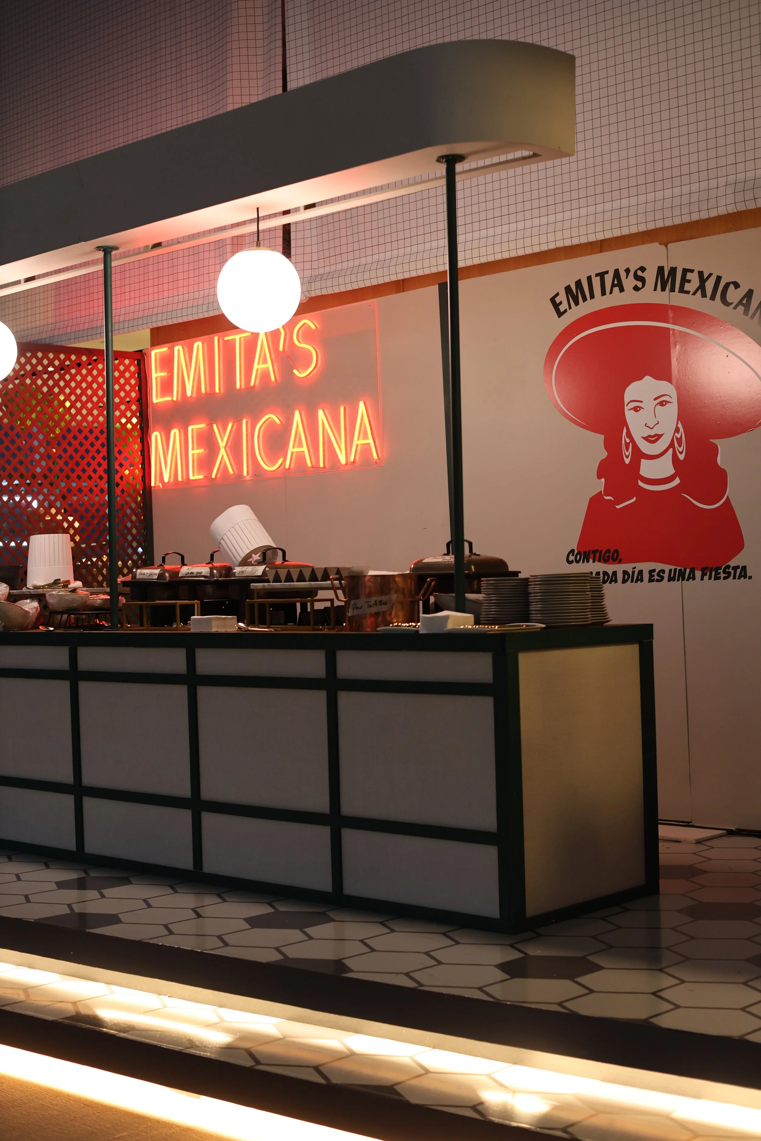 An interior of a Mexican restaurant with a neon sign that says 'Emita's Mexicana', a logo of a woman with a sombrero on the wall, and a buffet counter with plates and food.