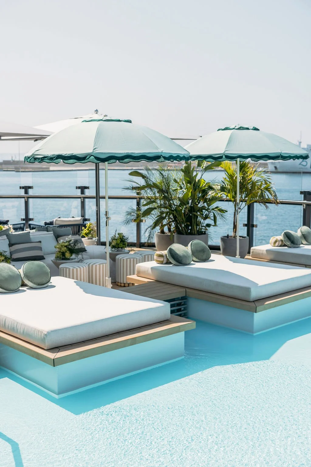 Luxury poolside lounge area with white cushioned sunbeds, green umbrellas, and potted plants overlooking the water and ships in the distance.