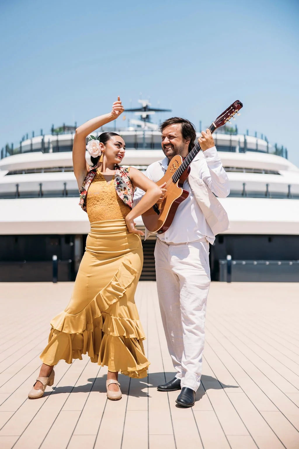A man and woman dance and sing on a ship's deck, with the man playing an acoustic guitar. The woman wears a yellow flamenco-style dress with ruffles, and the man wears a white outfit. The ship's structure is visible in the background under a clear bl