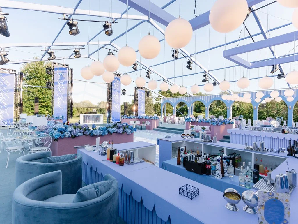 Indoor event space decorated in pastel blue and pink with hanging round paper lanterns, floral arrangements, bar area, and tables for a celebration or party.