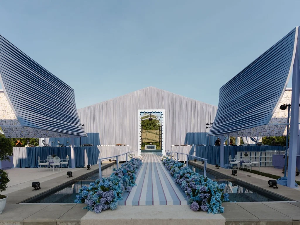Elegant outdoor event space with a striped aisle rug, surrounded by blue and white flowers, and modern structures with metallic and fabric accents under a clear sky.