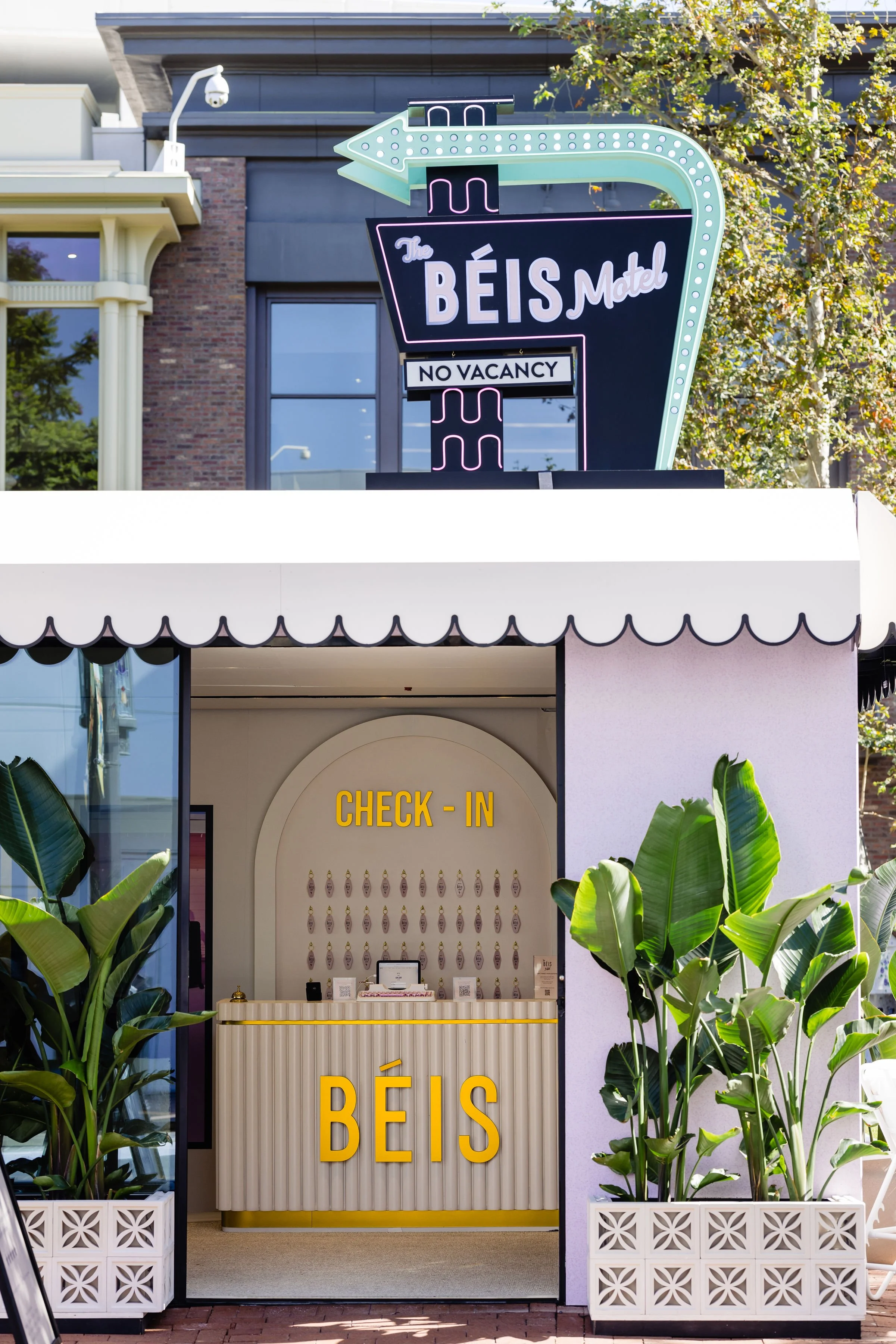Exterior of a motel with a neon sign reading 'The Beis Motel' and 'No Vacancy.' The entrance has a check-in counter with a sign that says 'Check-In' and 'BÉIS.' There are green plants on either side of the entrance.