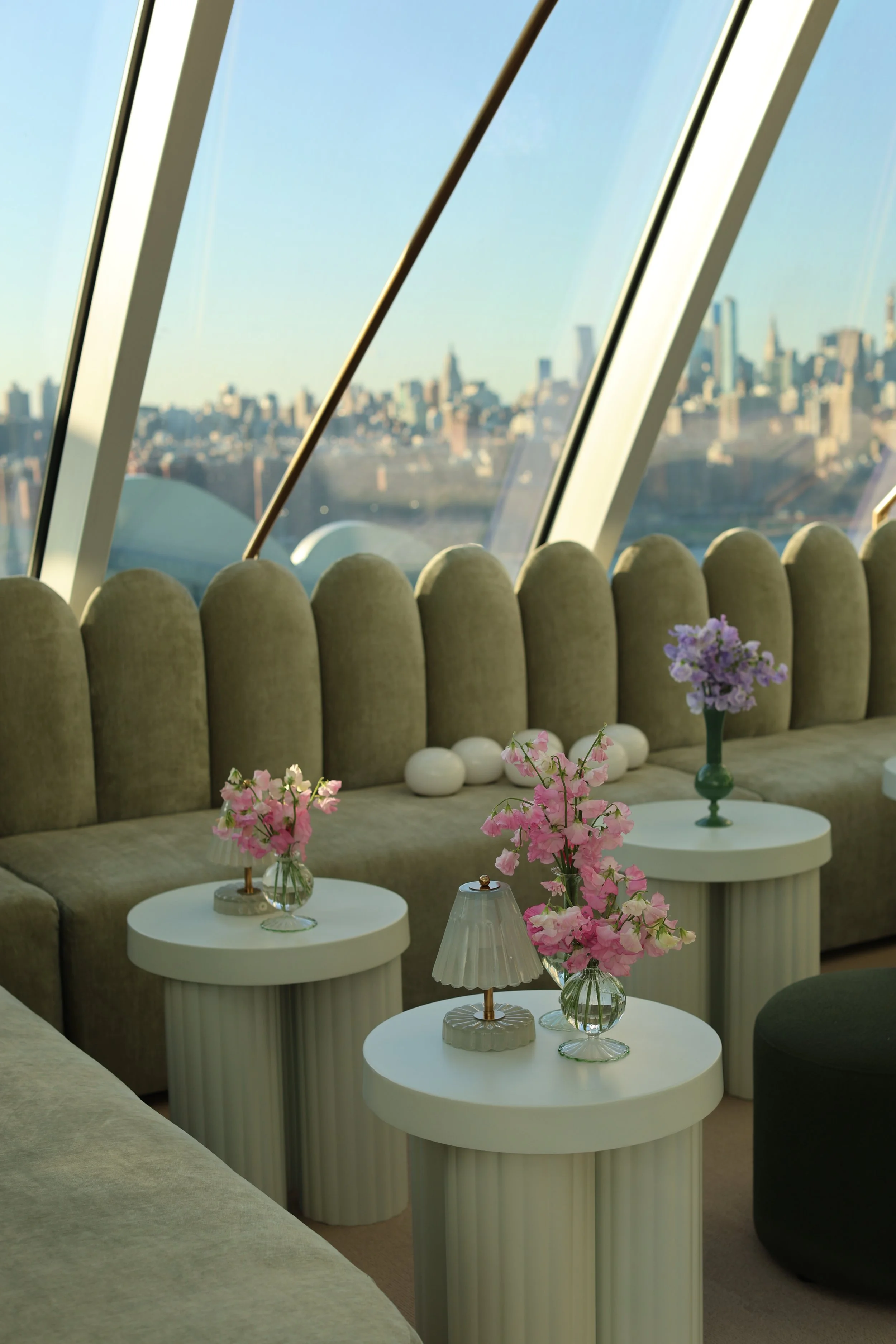 Interior of a modern lounge with a large curved window showing a city skyline. There is a long beige curved sofa, three white oval side tables with decorative lamps and pink and purple flower vases.