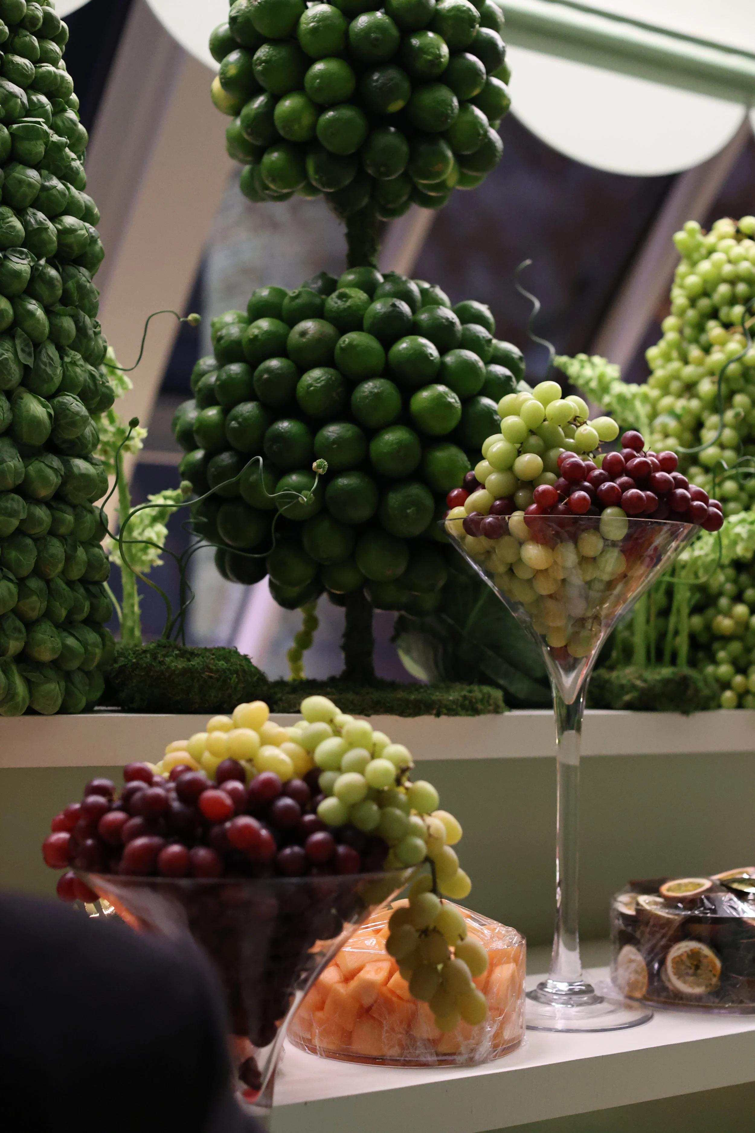 Decorative fruits including green and red grapes, arranged on a display with some in glass bowls and others on faux trees.