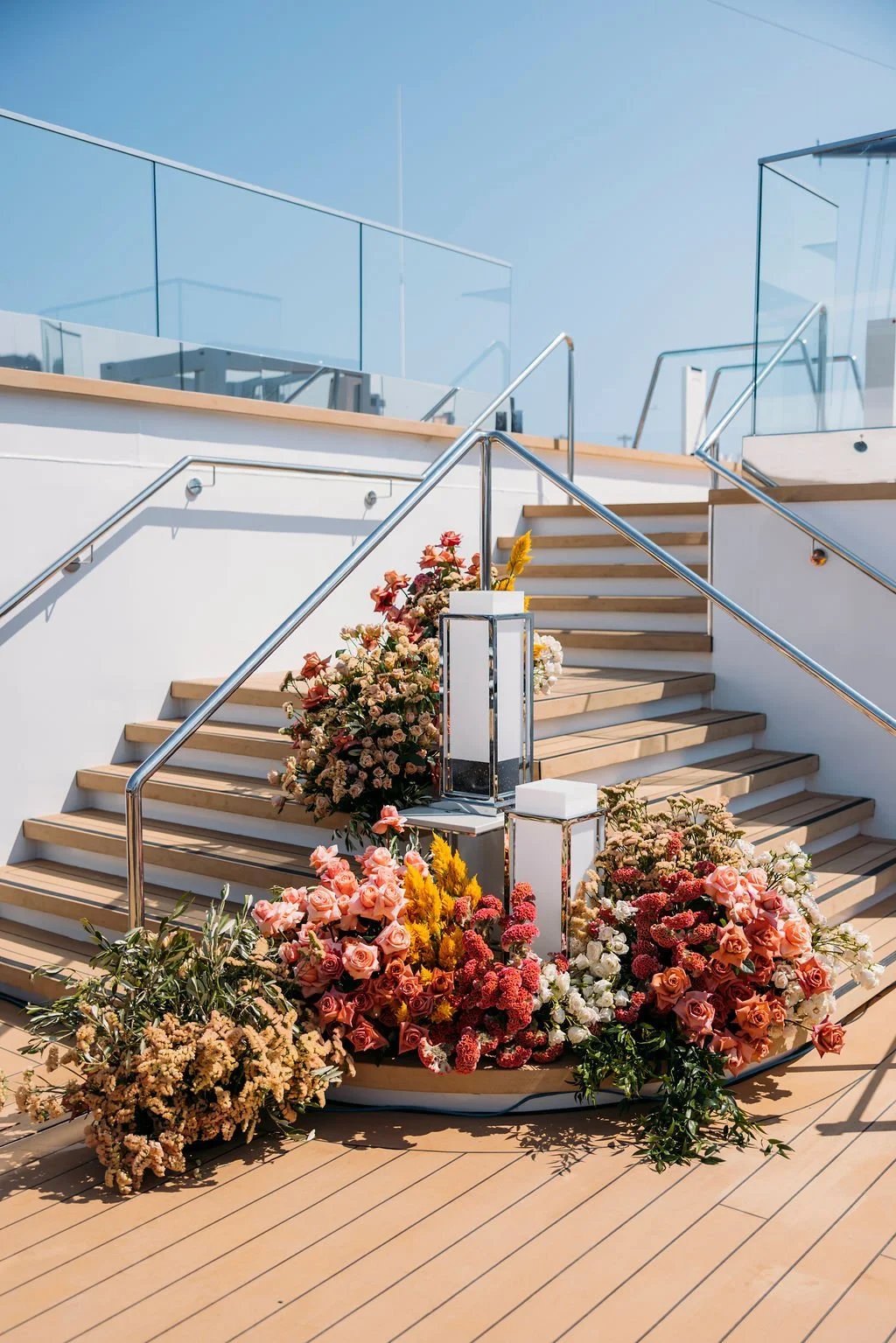 Elegant outdoor staircase decorated with colorful flowers, leading to a rooftop terrace with glass railings and clear blue sky.
