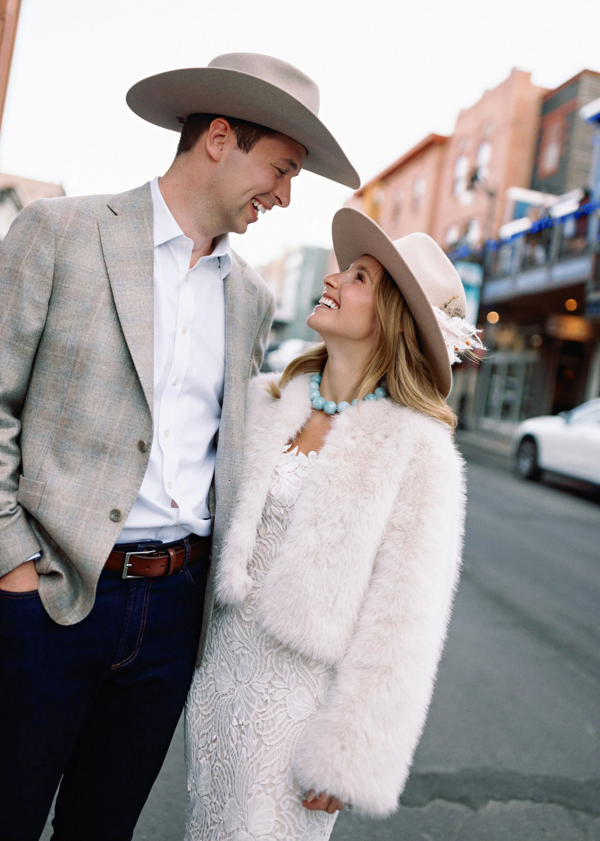 A man and woman smiling and looking at each other on a city street, wearing stylish clothing and wide-brimmed hats.