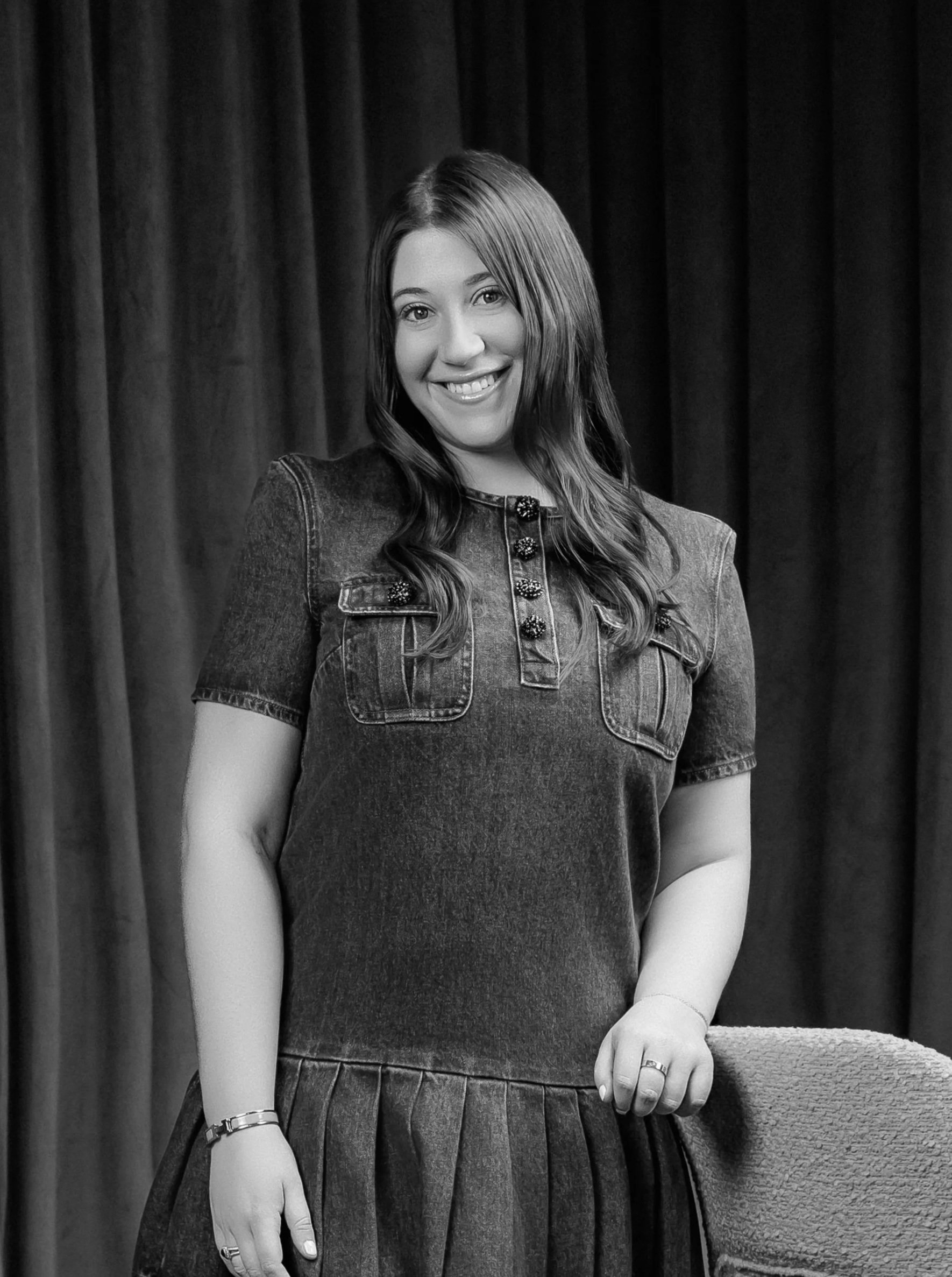 A smiling woman with long wavy hair wearing a short-sleeved denim dress, standing in front of dark curtains.