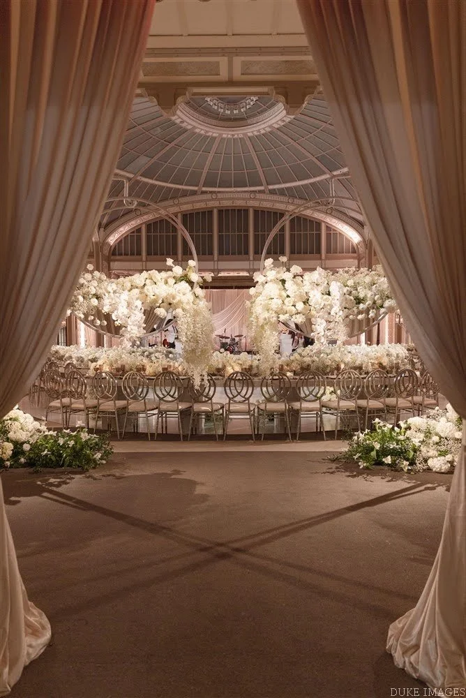 Elegant wedding reception setup with a floral centerpiece, round tables, and ornate chairs inside a glass-domed conservatory.