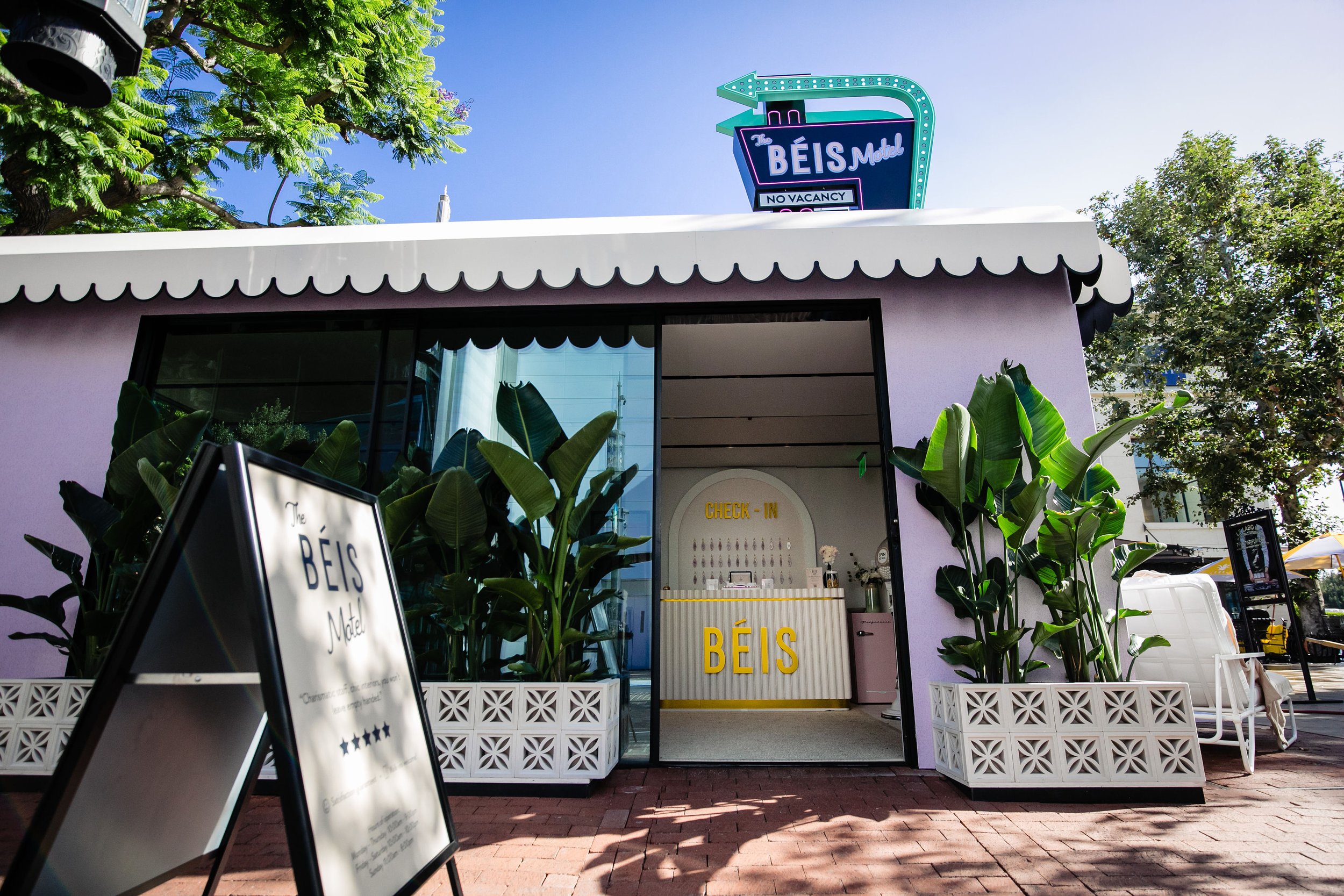 The exterior of a pastel purple building with a white scalloped awning, large potted plants, and a sign showing the name 'The Bêis Motel'. There is also a black sandwich board sign outside, and a check-in counter with the label 'Bêis' inside.