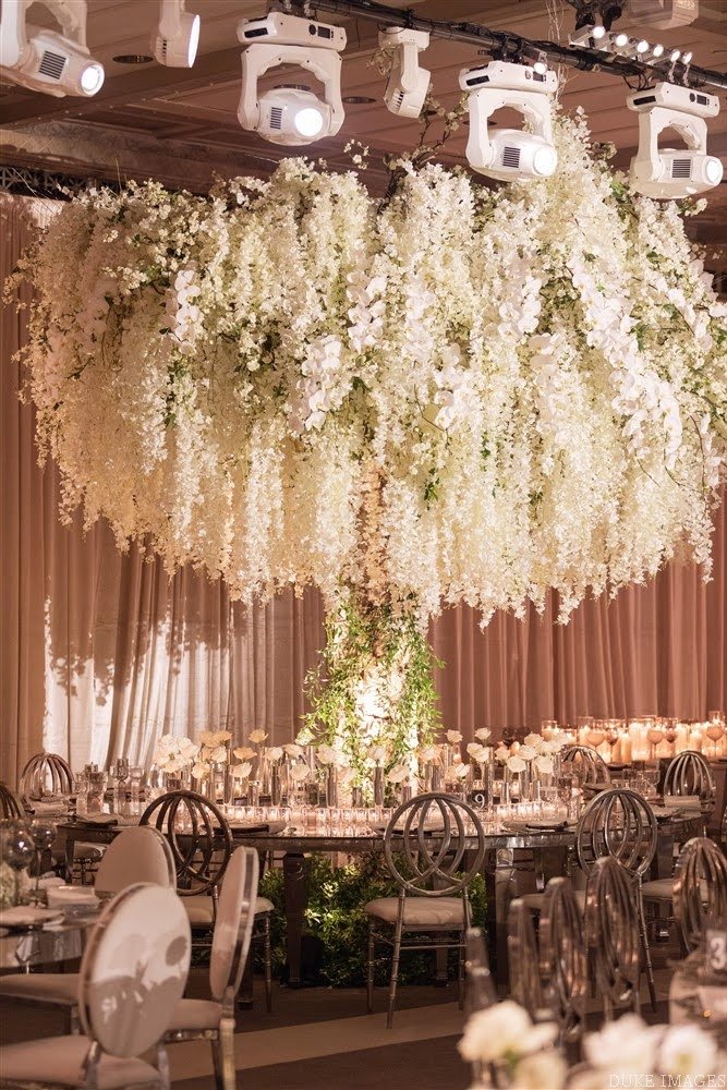 An elegant banquet hall decorated with a large floral chandelier and beige curtains, with round tables set for a formal event.