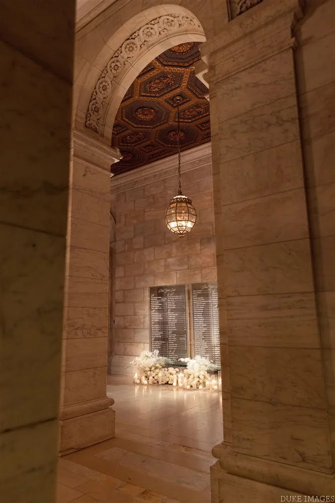 View of memorial plaques surrounded by candles and flowers inside a grand, marble hall with ornate ceiling and hanging lantern, possibly in a religious or historic building.