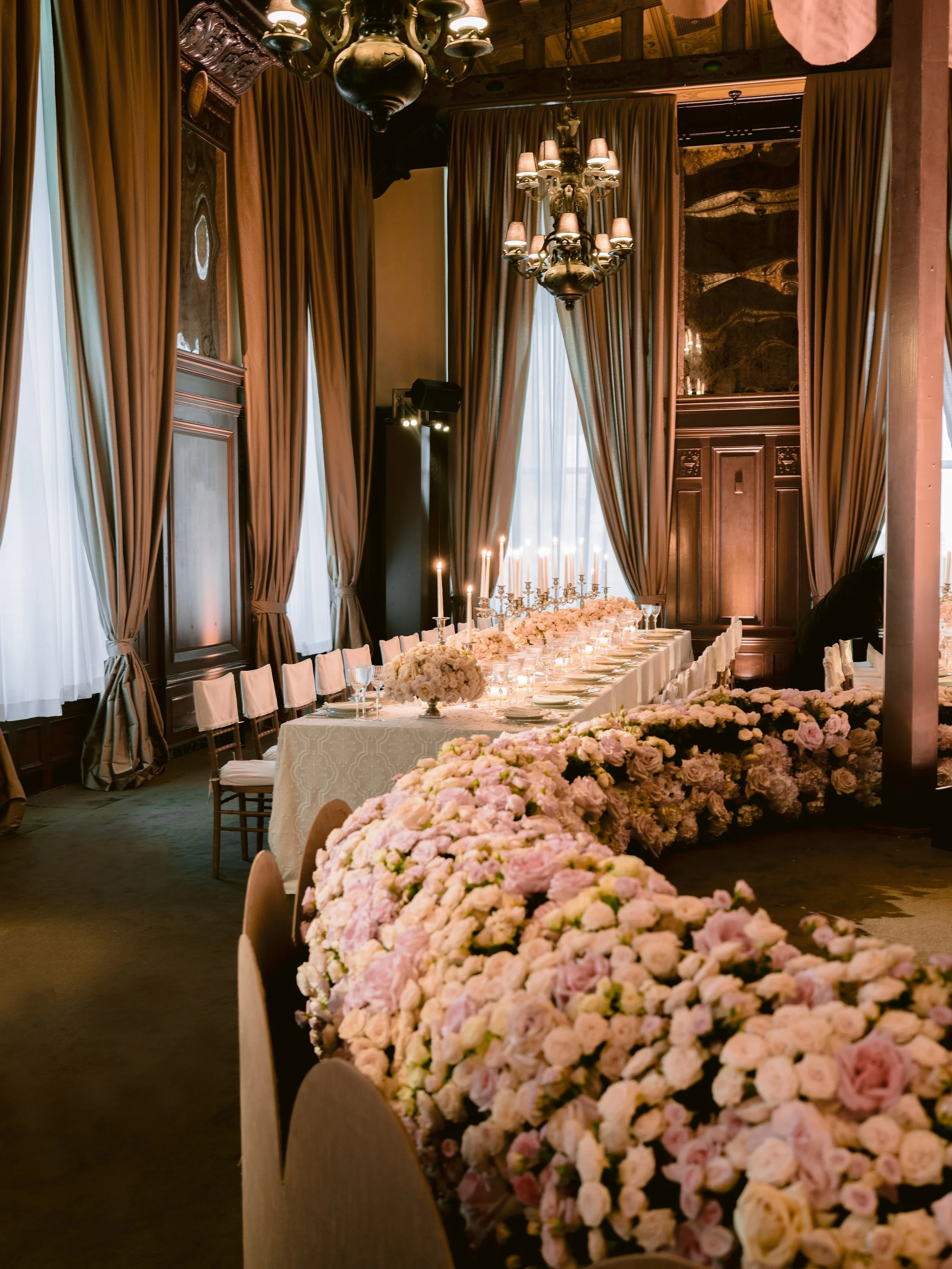 Elegant banquet room with a long table decorated with pink and white flowers, tall candelabras with lit candles, and surrounded by wooden chairs, with large curtains, chandeliers, and wooden wall paneling.