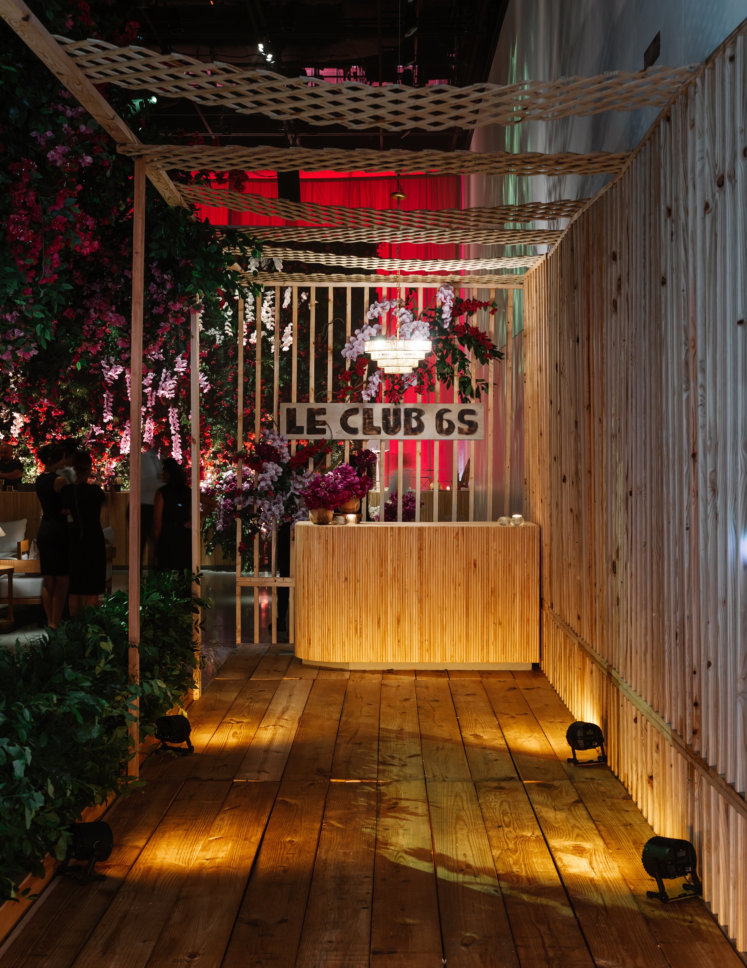 An event space decorated with pink and white flowers, wooden walls and flooring, and a sign that reads 'LE CLUB 6S'. There is a wooden reception desk and a chandelier overhead.