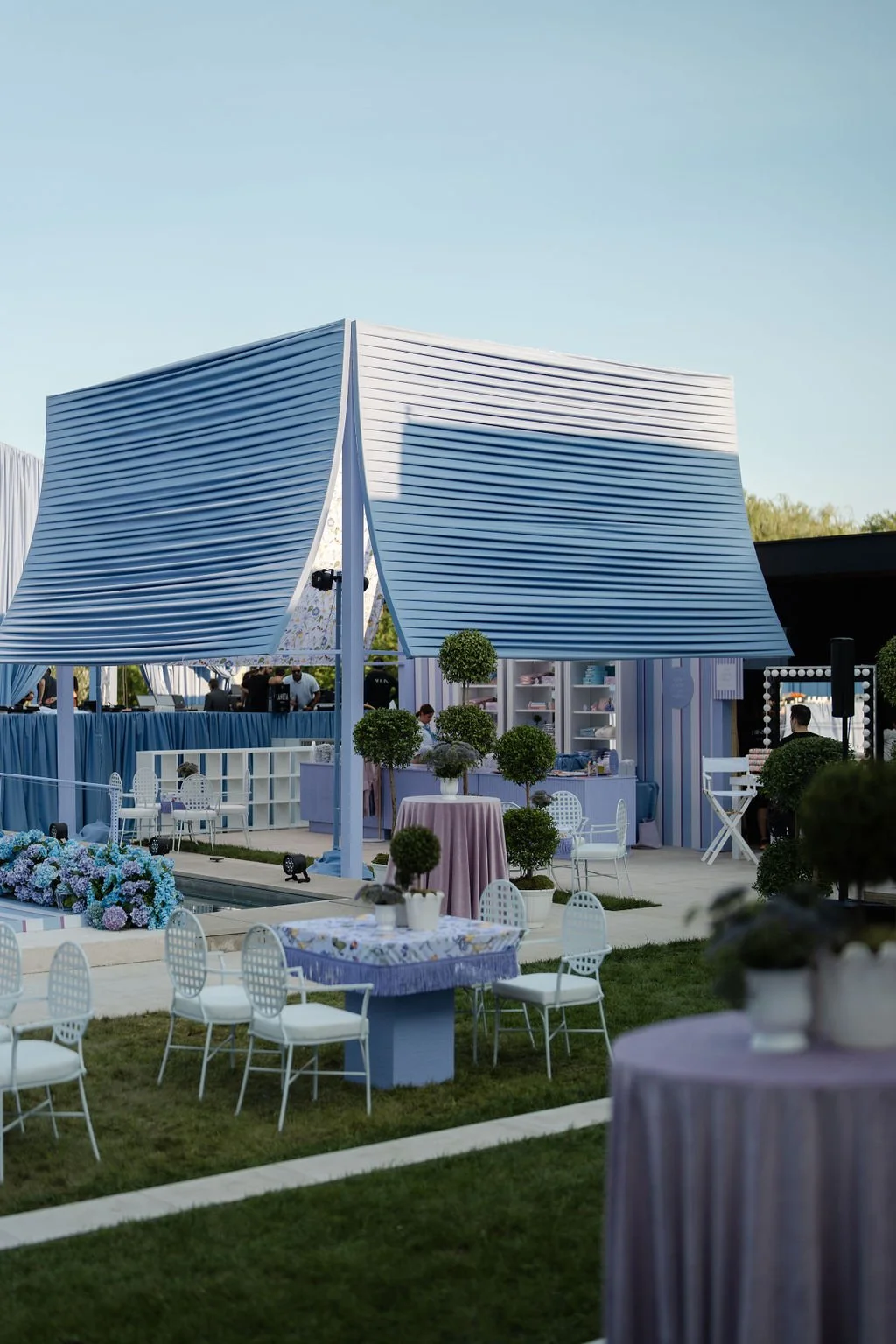 Outdoor event setup with a blue and white color theme, featuring a striped canopy, decorated tables with flowers, potted bushes, and chairs, with a clear sky in the background.