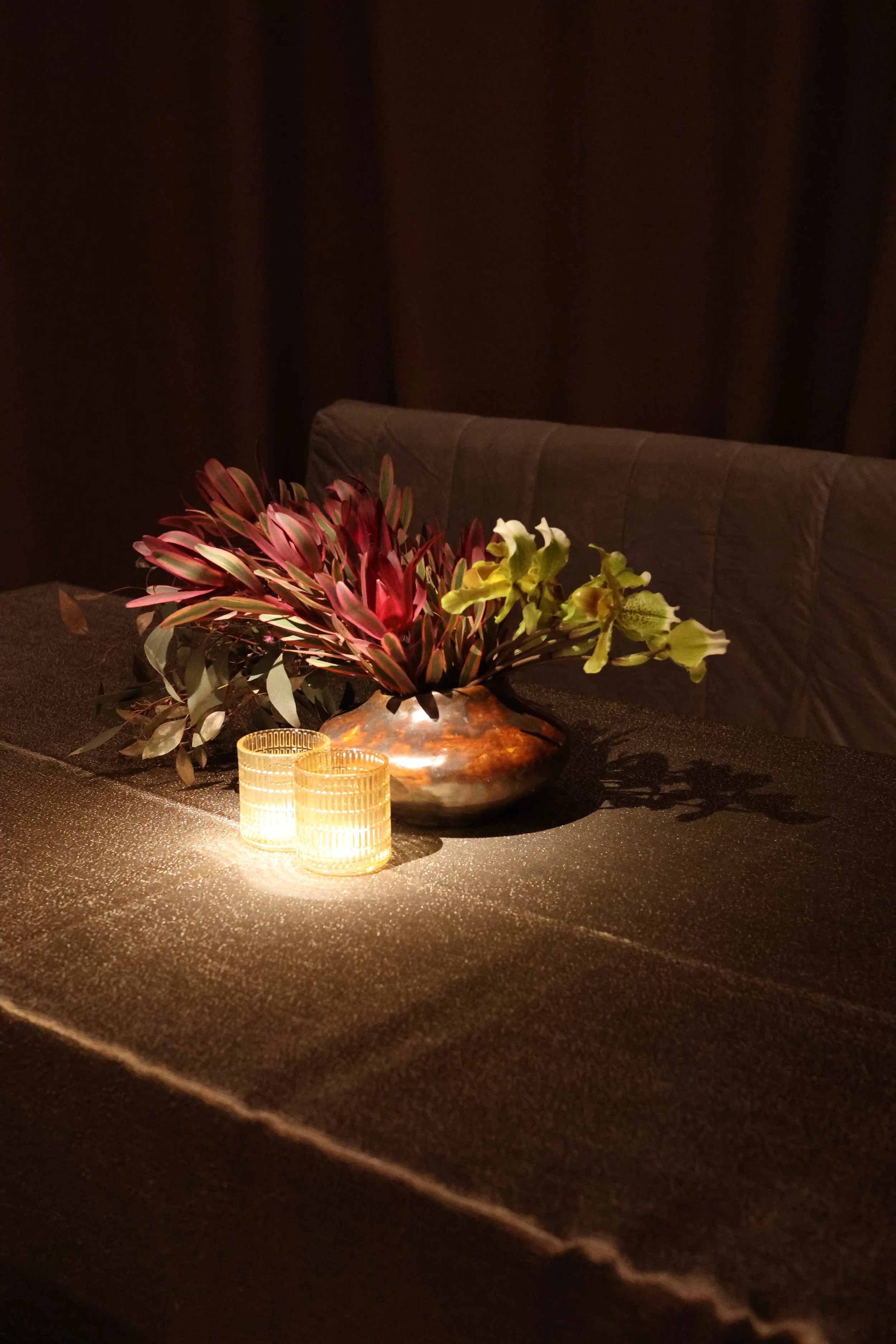 A dark dining room table with a floral centerpiece and two small lit candles, with a dark upholstered headboard in the background.