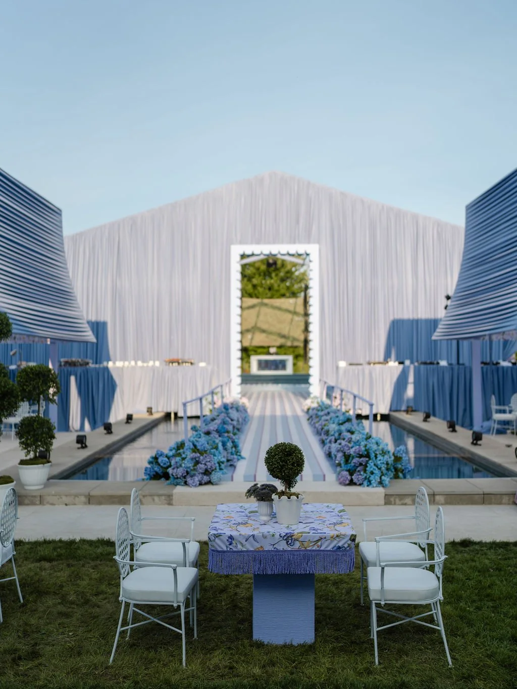 Elegant outdoor event space with a white fabric-draped structure, floral decorations, and a central aisle leading to an altar, with white chairs and tables outside on a grassy area.