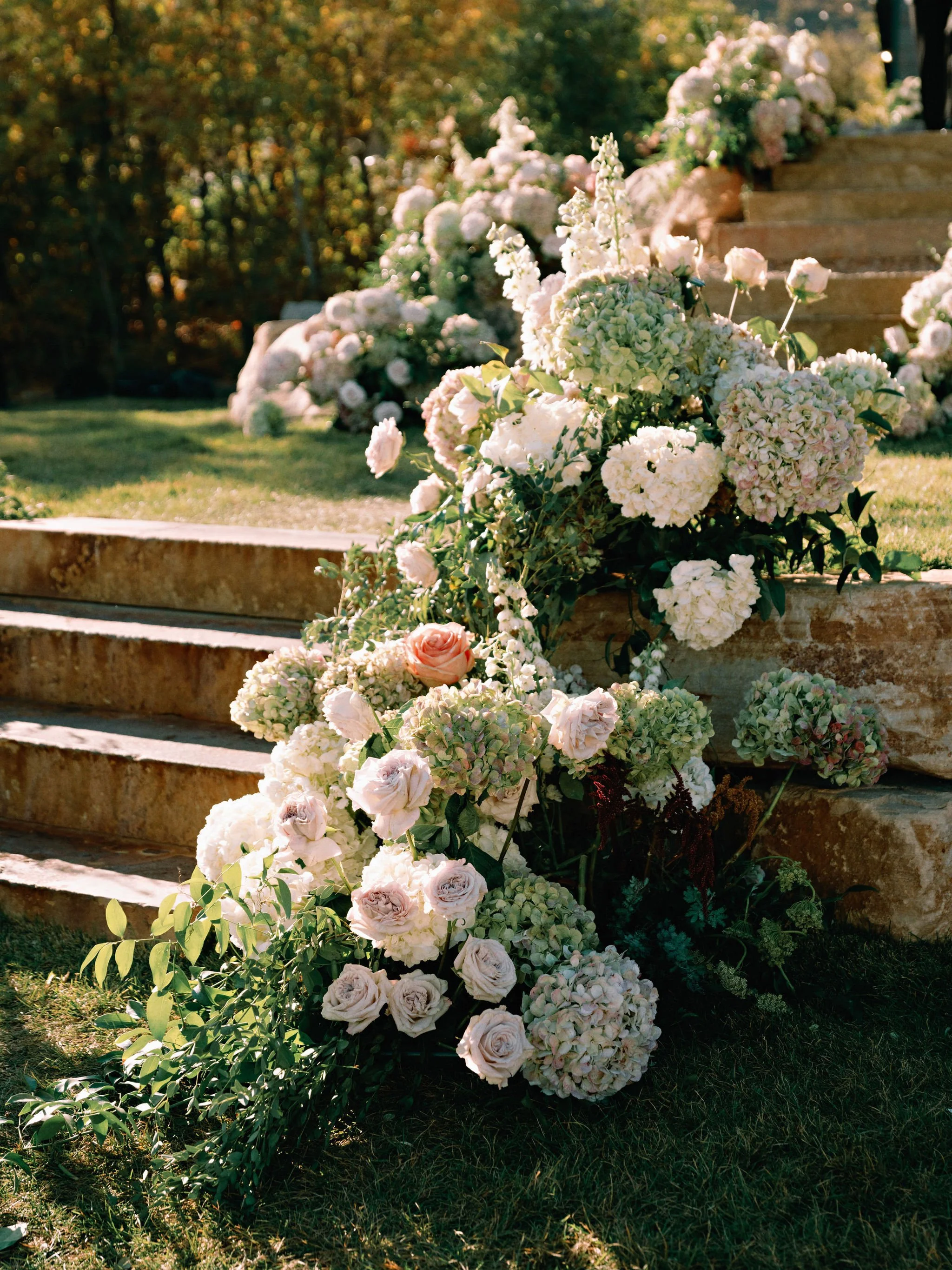 Arrangement of pink and white flowers, including roses and hydrangeas, on stone steps outdoors with greenery in the background.