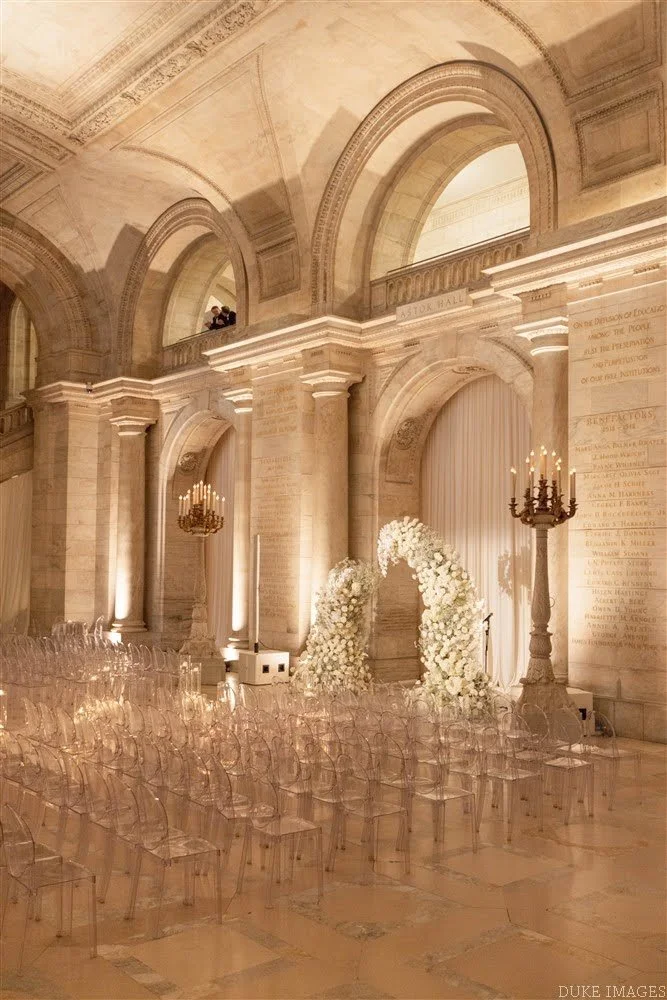Elegant indoor wedding ceremony setup with floral arch, transparent chairs, chandeliers, and soft lighting inside a grand historic building.