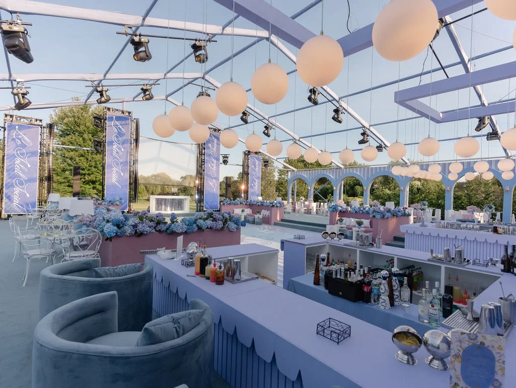 Wedding reception area with pastel pink and blue decor, floral arrangements, balloon ceiling, stage with LED screens, and a bar area with various bottles and glassware.