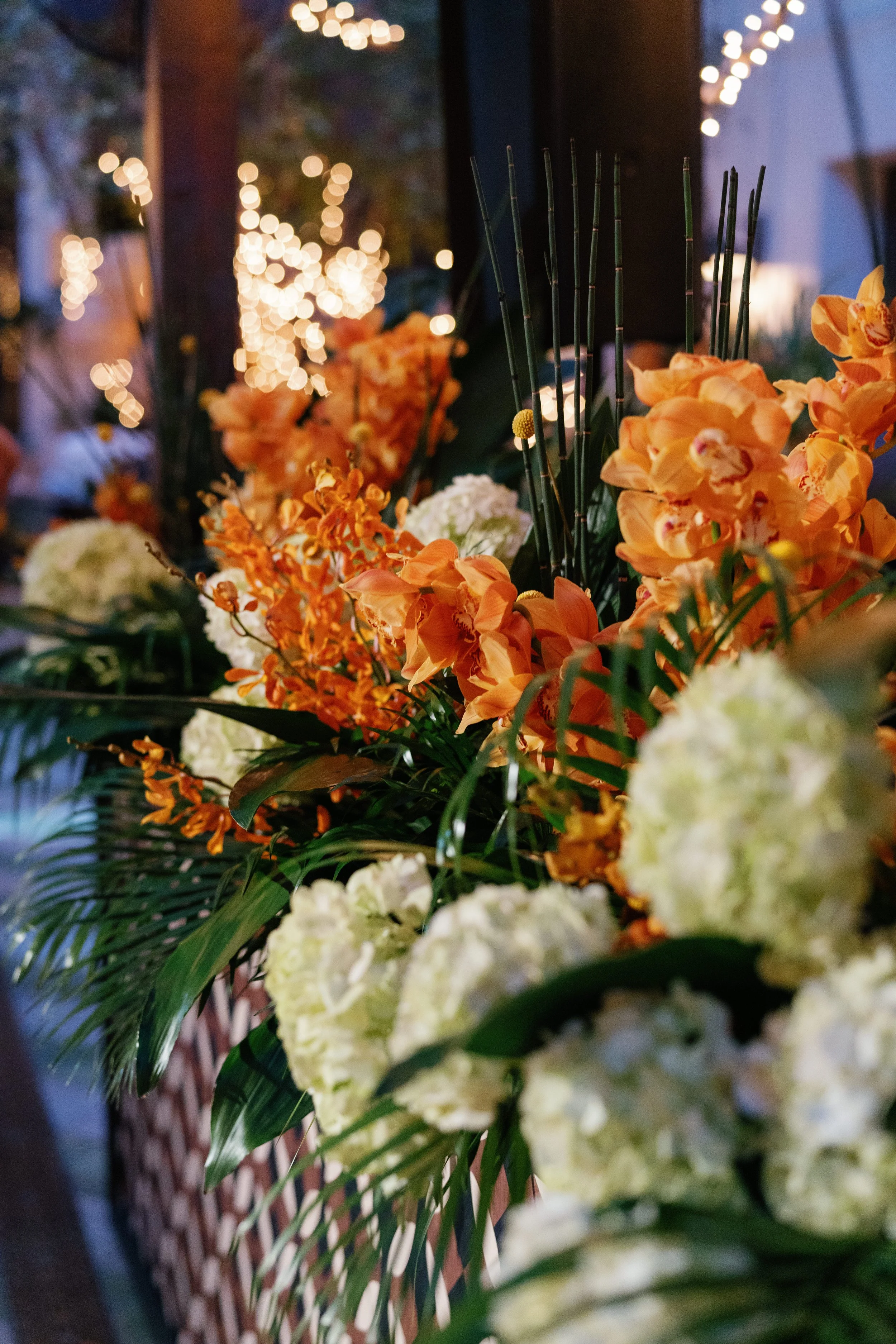 Close-up of floral arrangement with orange and white flowers, blurred string lights in the background.