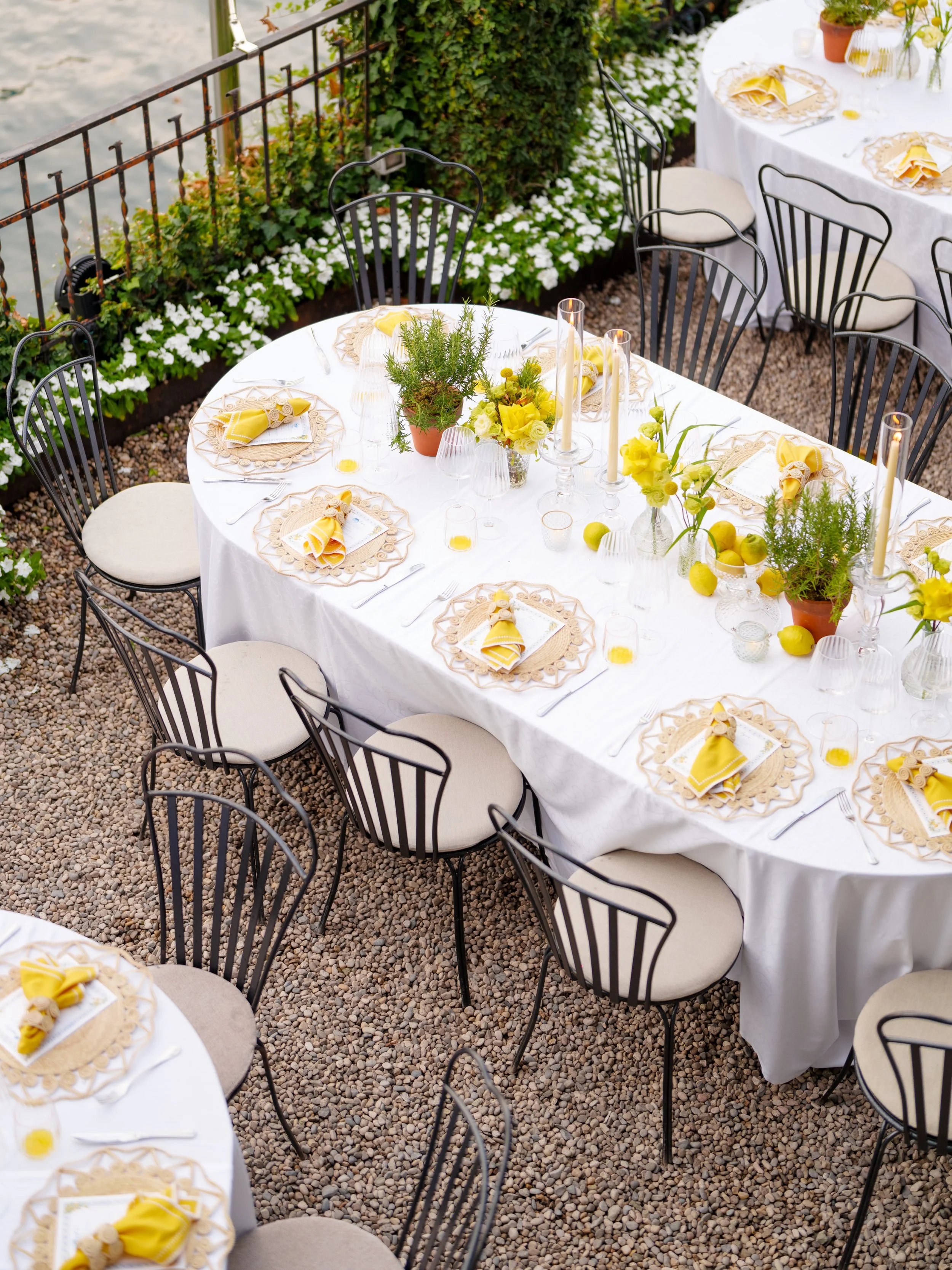 Round outdoor dining table with a white tablecloth, yellow napkins, floral centerpieces, candles, and place settings, surrounded by black chairs on a gravel surface with potted plants and white flowers.