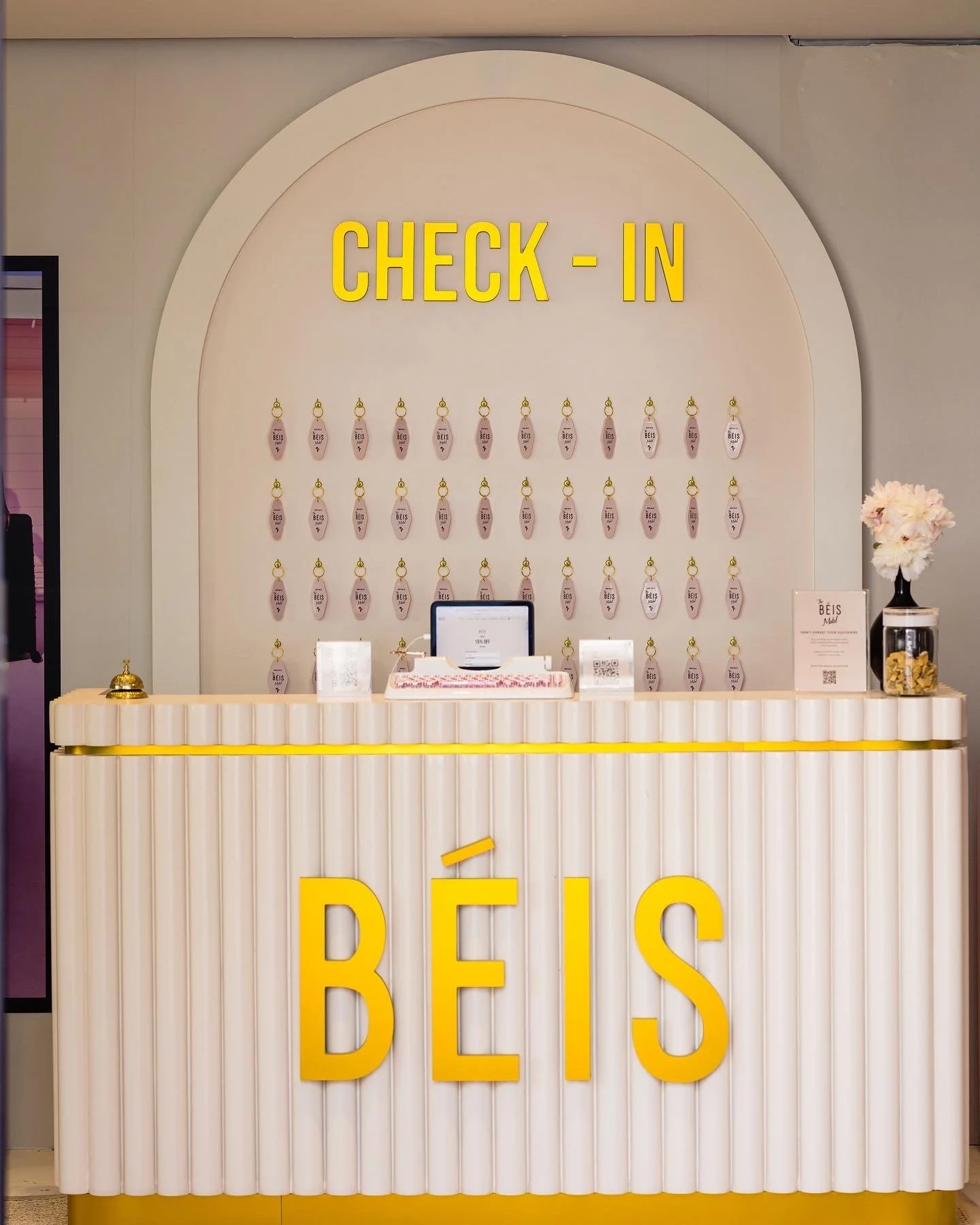 A check-in counter with a sign reading 'CHECK-IN' and hanging tags behind it. The counter has large yellow letters spelling 'BÉIS' on the front, with a computer, a bell, and informational signs on top.