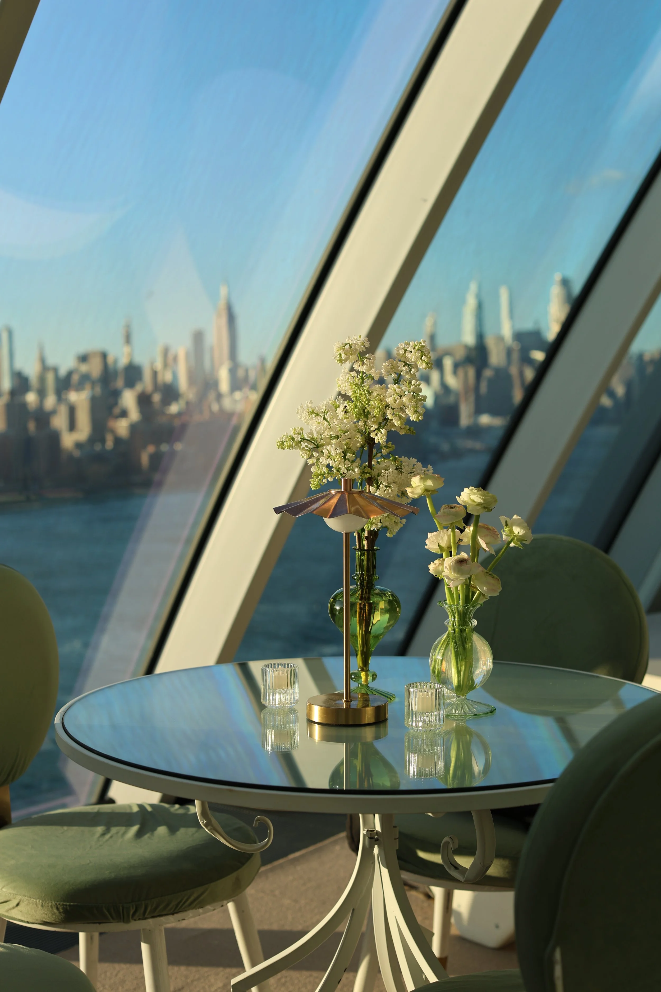 A round dining table with green chairs, floral centerpieces in vases, and a city skyline view outside a large window.