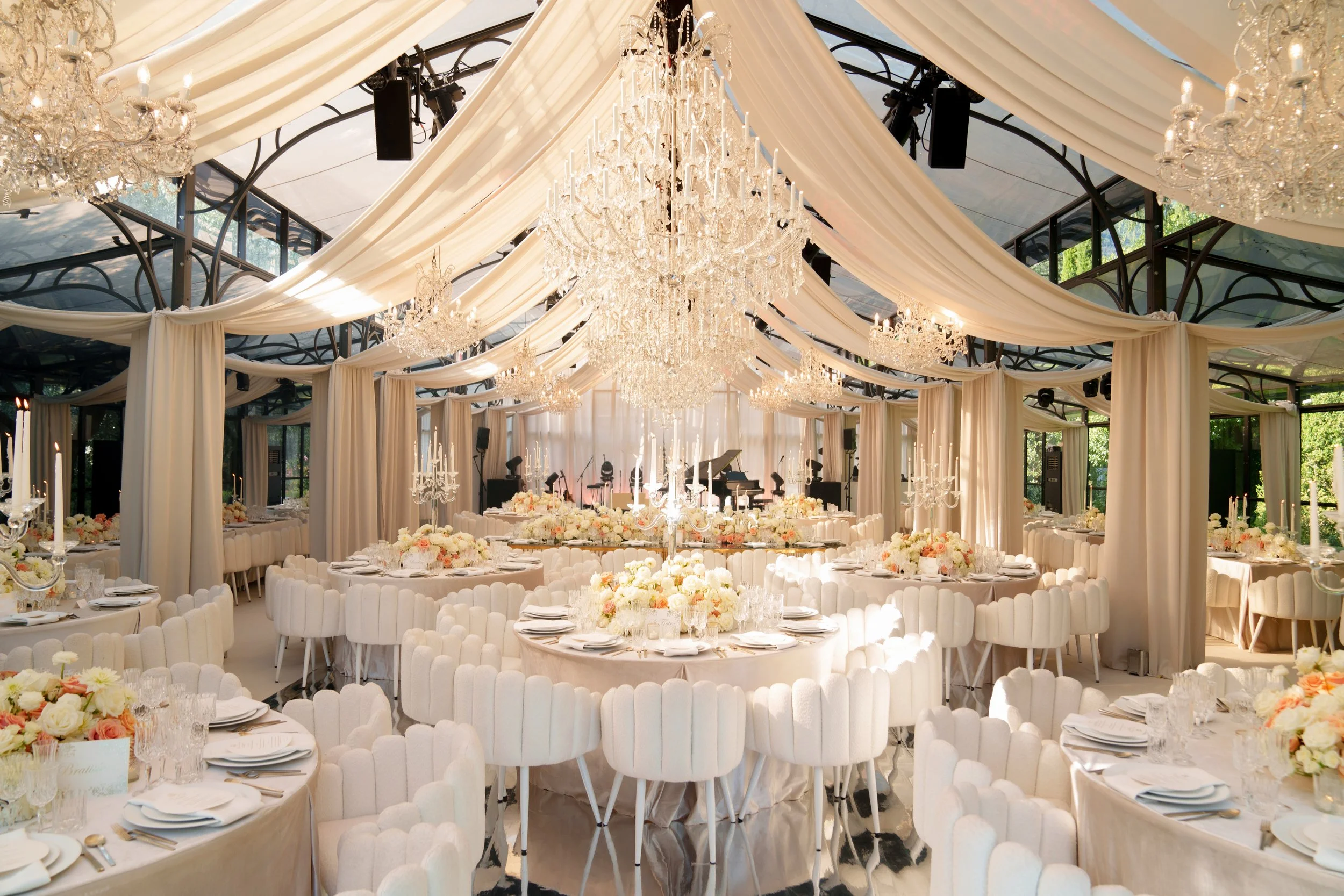 Elegant event space with white drapes, chandeliers, flower centerpieces, and tiered table arrangements, decorated for a wedding or celebration.