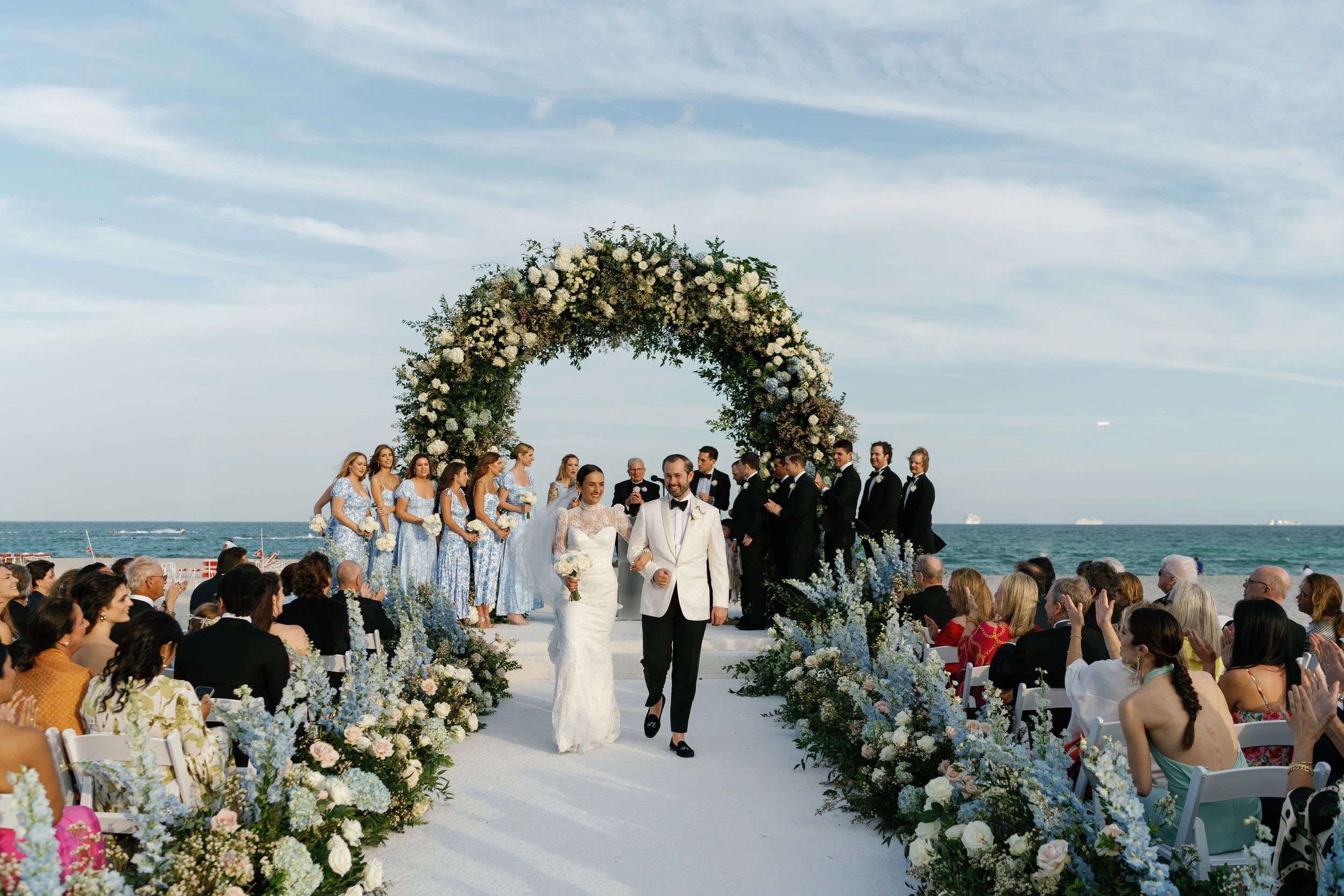 A wedding ceremony on the beach with a bride and groom walking down the aisle, surrounded by guests, a floral arch, and the ocean in the background.