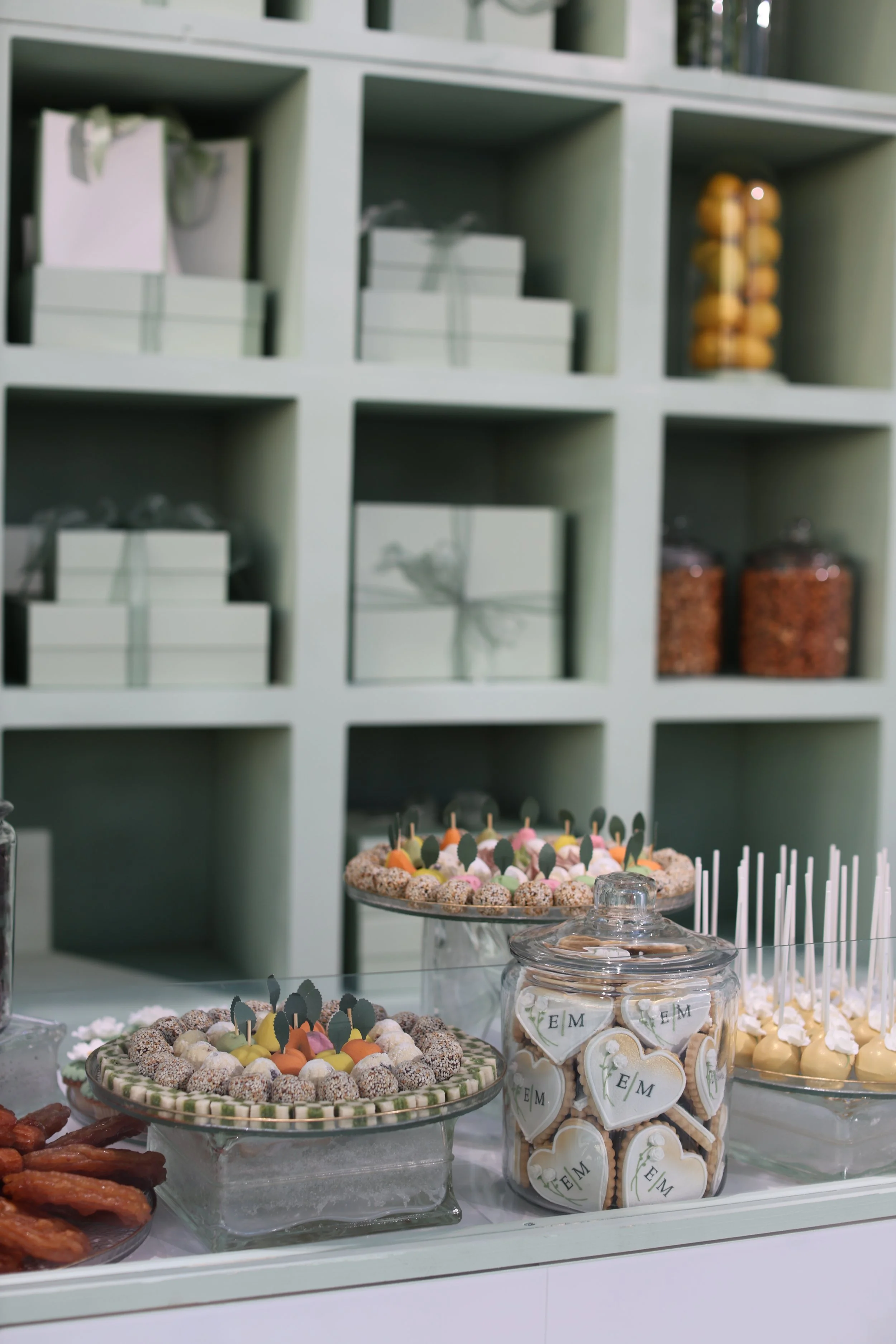 Dessert table with cookies, cake pops, and sweets in front of a wall unit with boxes and jars.