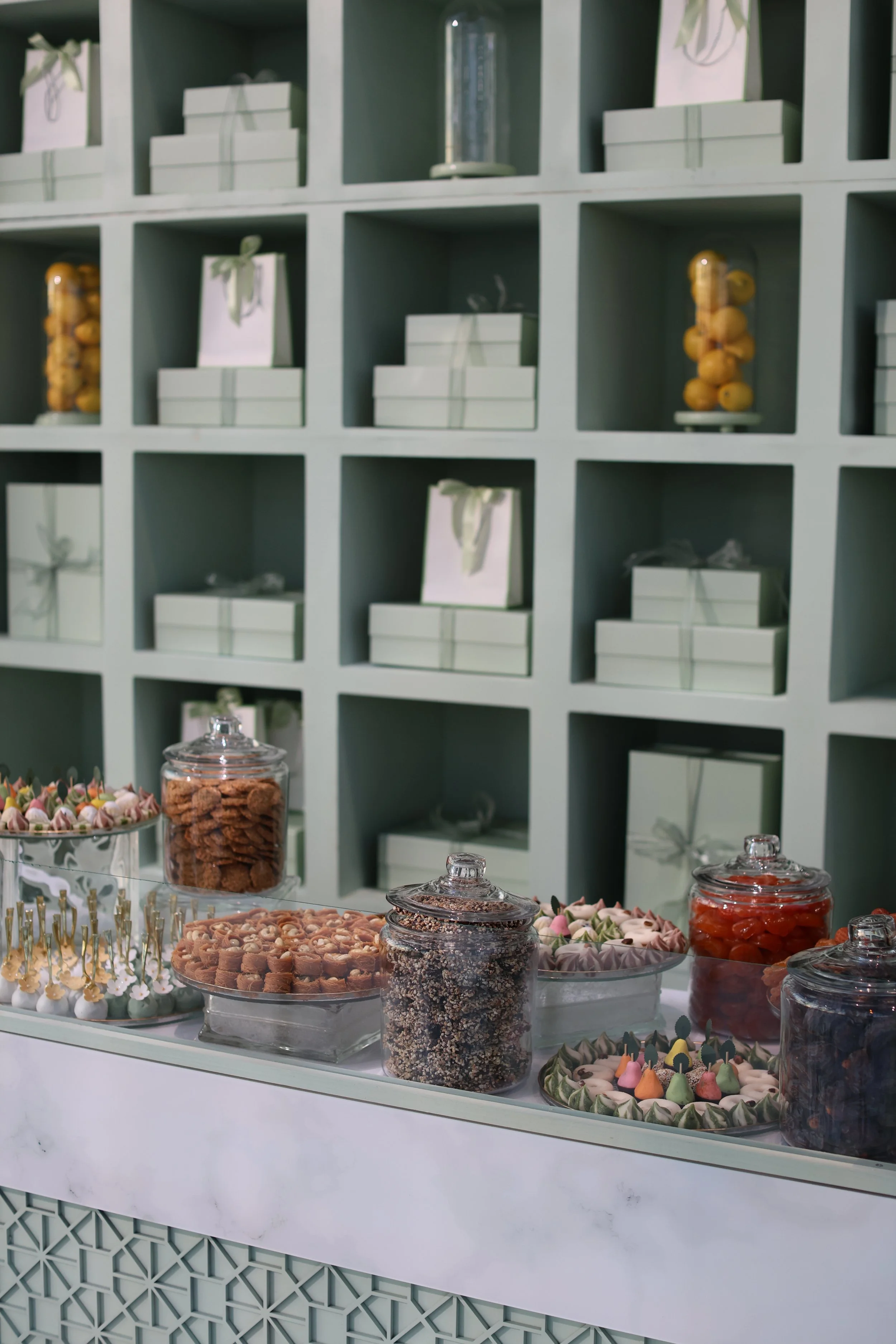 Display of sweets and candies on a table in front of a green shelving unit with gift boxes and decorative items.