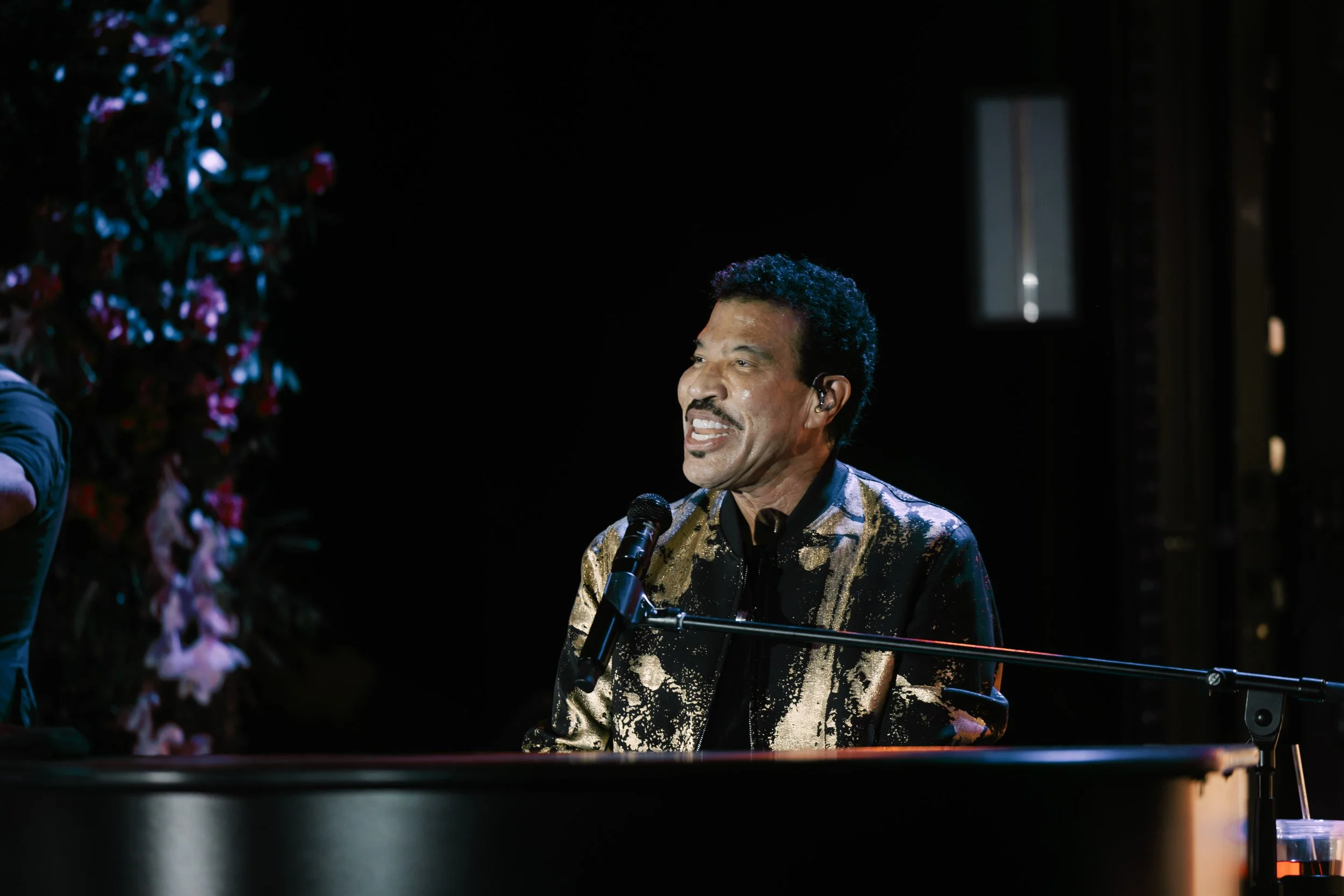 A man singing into a microphone while playing the piano, wearing a shiny black and gold jacket on a dark stage.