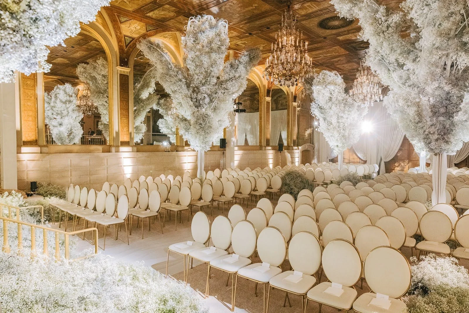 Elegant wedding or event venue decorated with white flowers, tall floral arrangements resembling trees, luxurious chandeliers, and a large seating area with rows of white chairs.