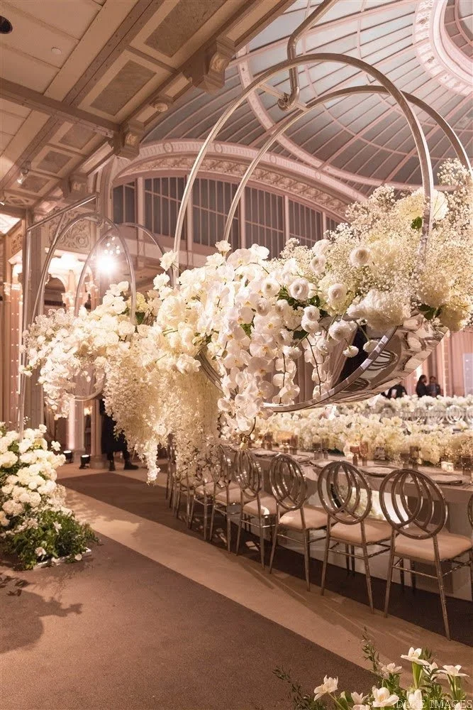 Elegant event space decorated with white flowers and floral arrangements, featuring hanging circular metallic structures, a long table with chairs, and a luxurious architectural ceiling.