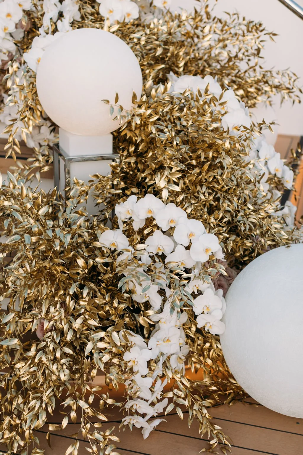 A decorative arrangement of white orchids, gold-colored leaves, and white spherical lamps.