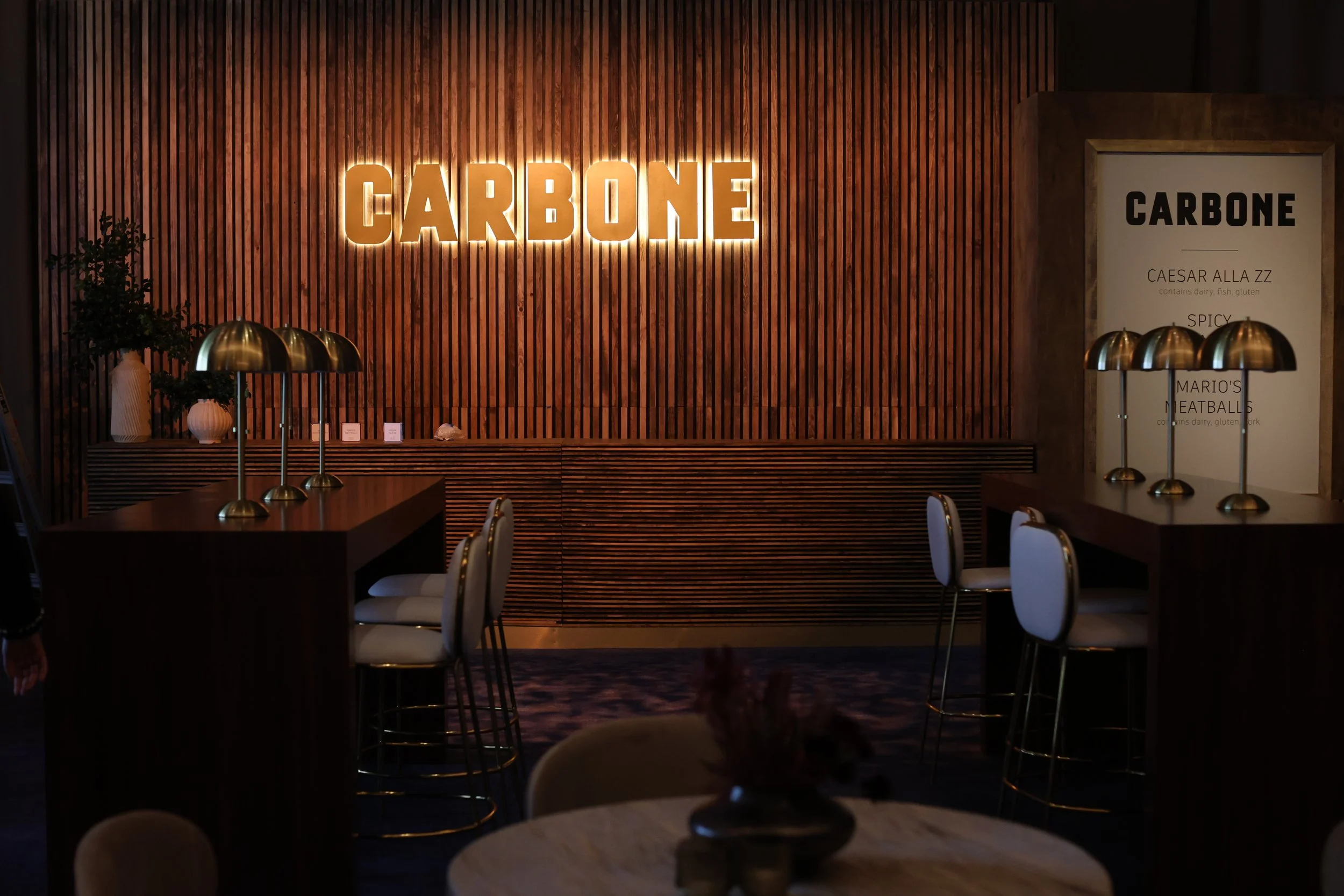 Interior of a restaurant with a wooden wall, illuminated sign that reads 'CARBONE,' a framed menu, tables with gold lamps, and chairs with white upholstery.